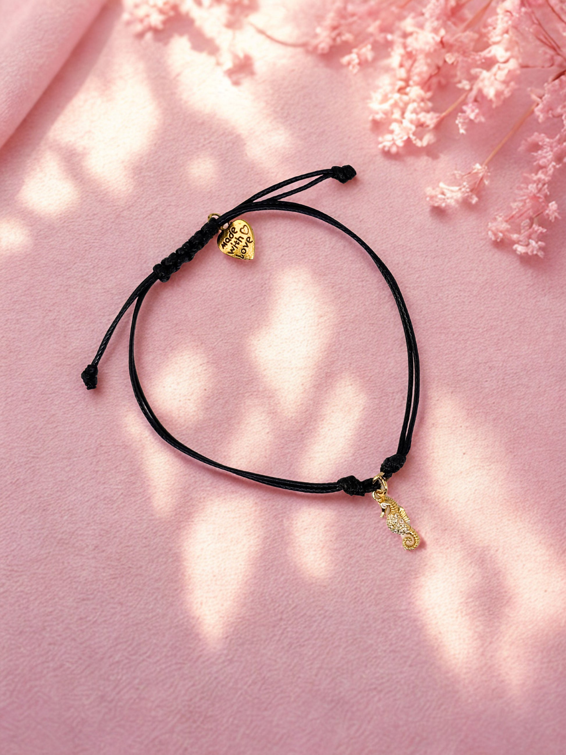 Black cord bracelet with gold seahorse charm on a pink background