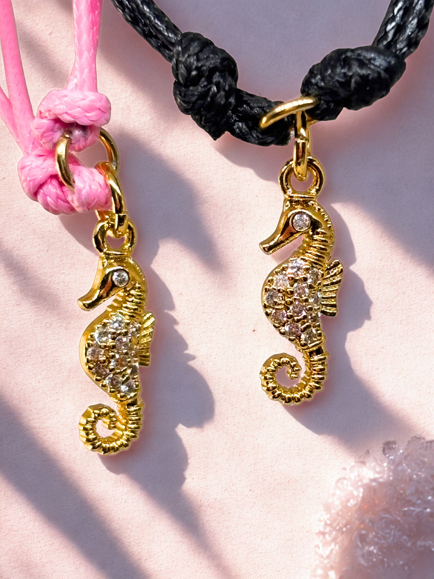 Two gold seahorse charms on pink and black cords against a pink background.