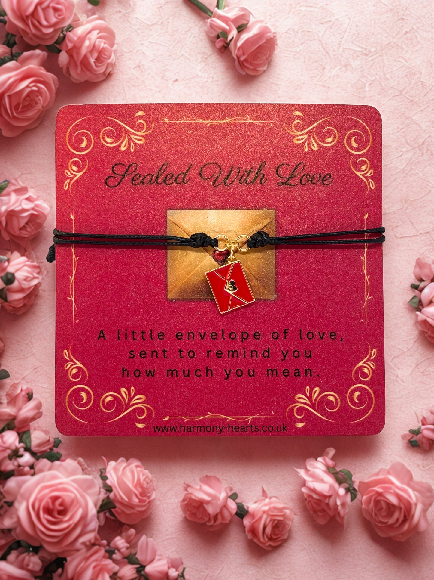 Red card with gold text 'Sealed With Love' and a charm bracelet with envelope charm on a pink background with pink roses.