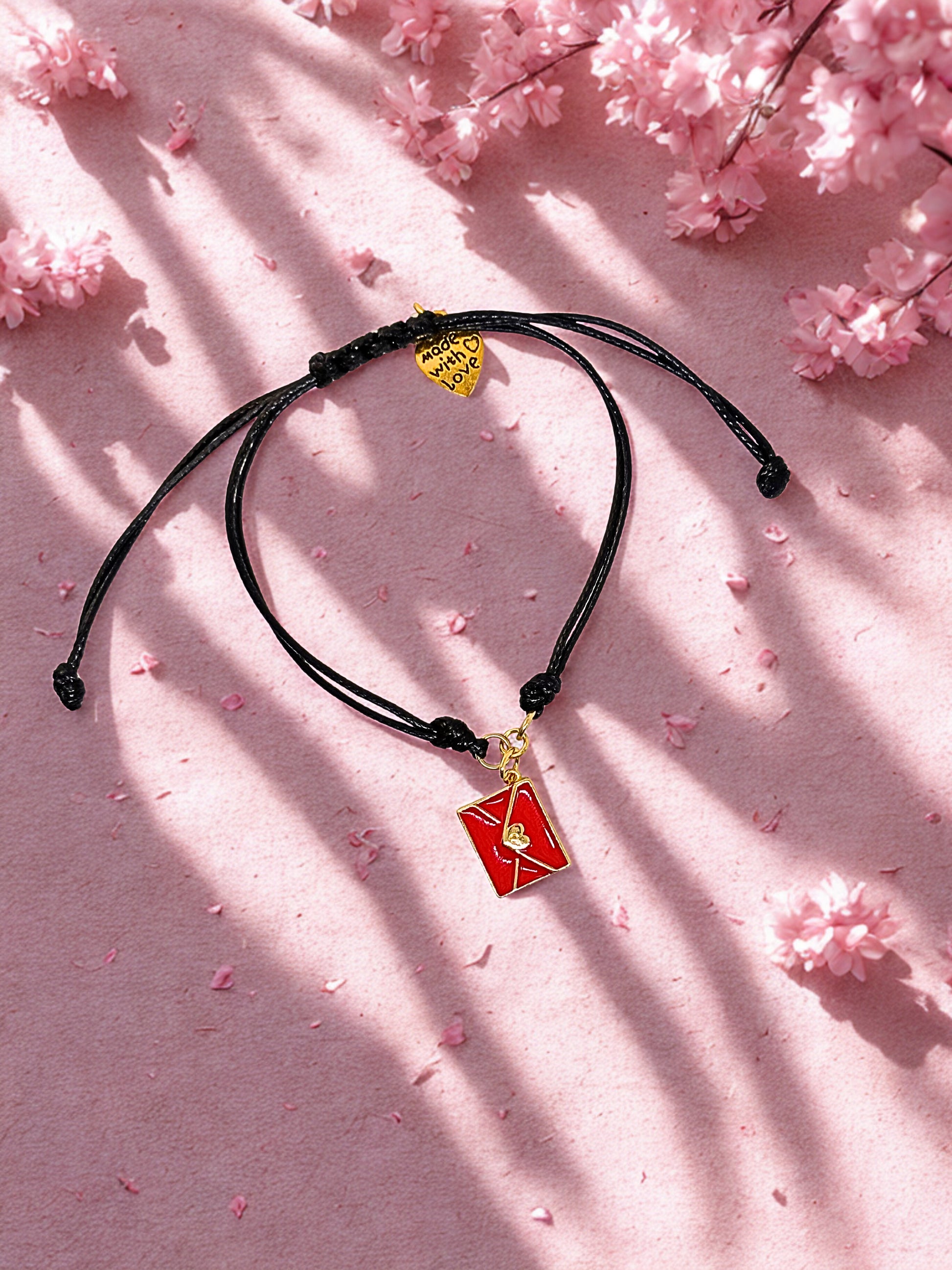 Black cord bracelet with red and gold envelope charms on a pink background with cherry blossoms