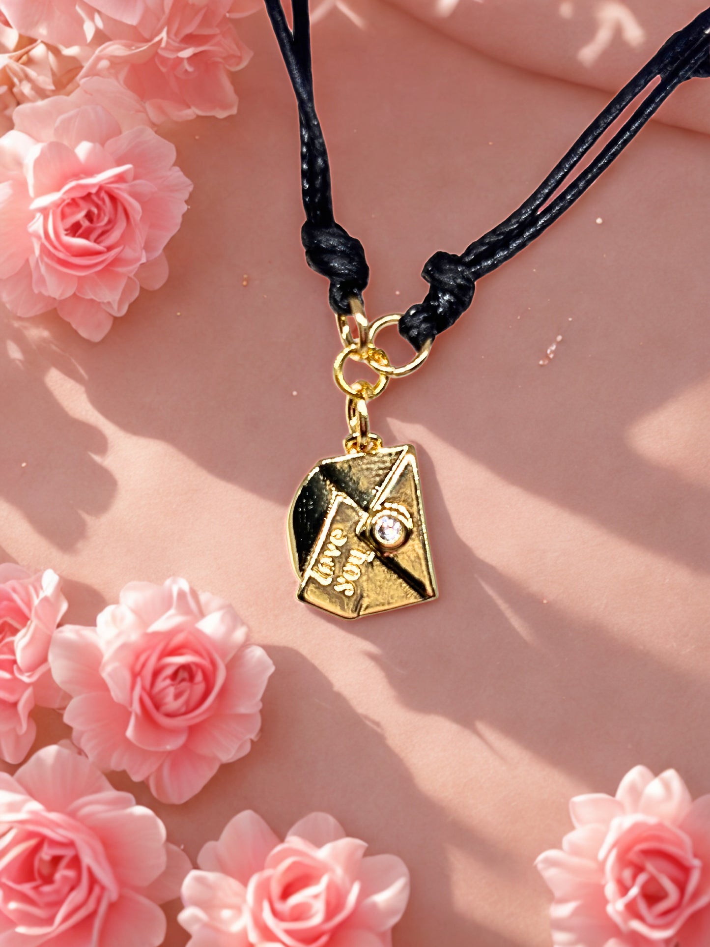 Gold envelope charm bracelet with a black cord on a pink floral background