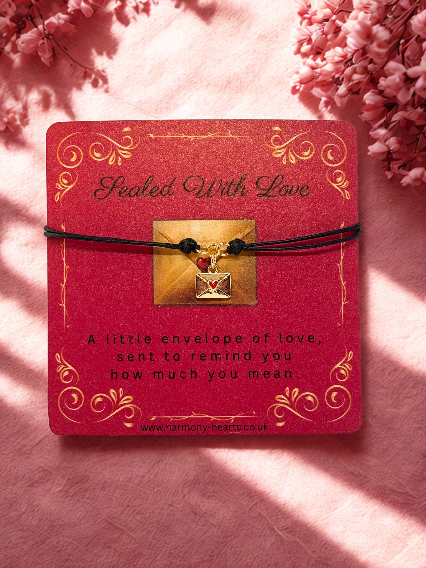 Red card with gold text and a small gold envelope charm bracelet on a pink background