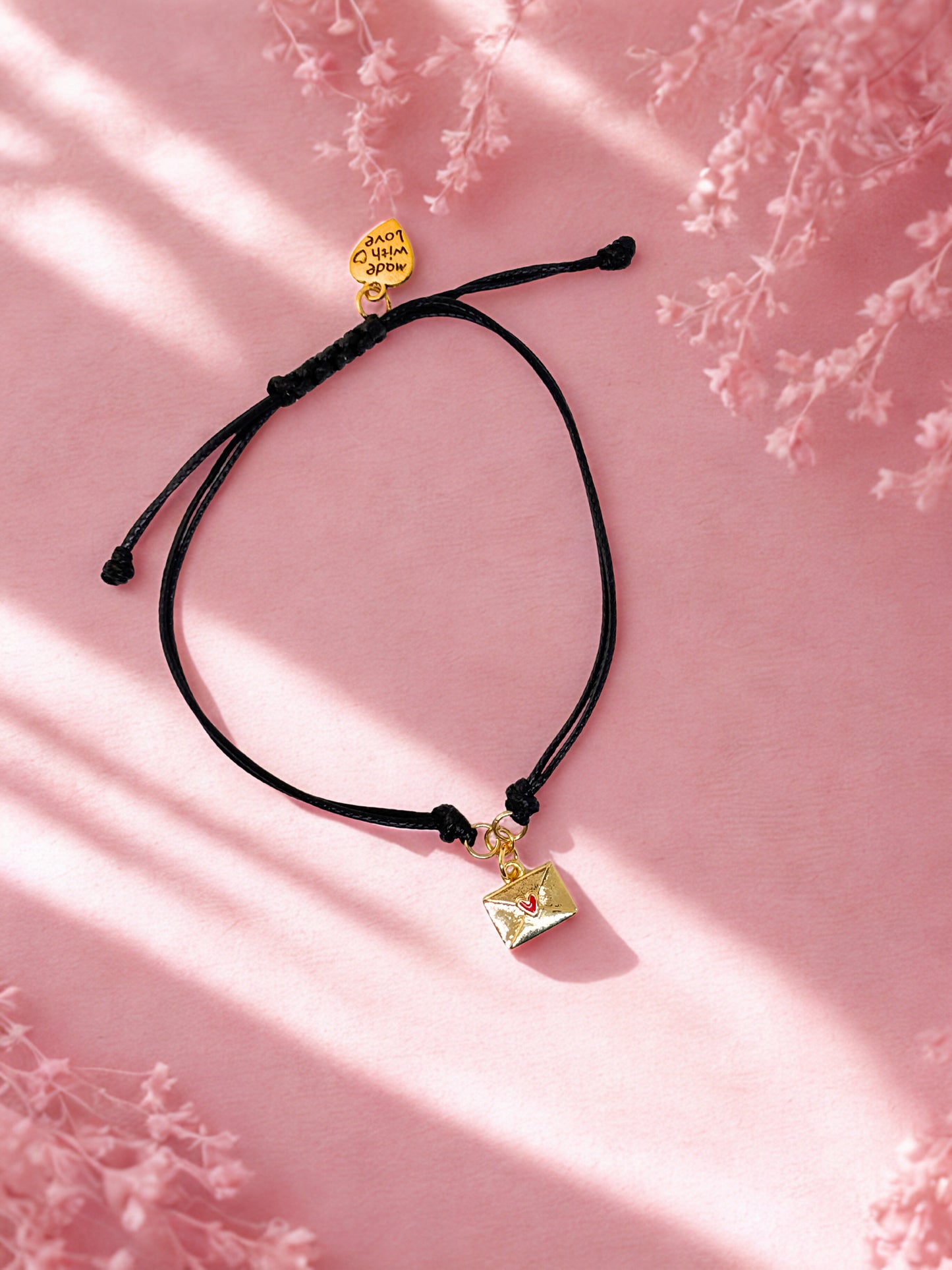 Black cord bracelet with gold envelope charm on a pink background