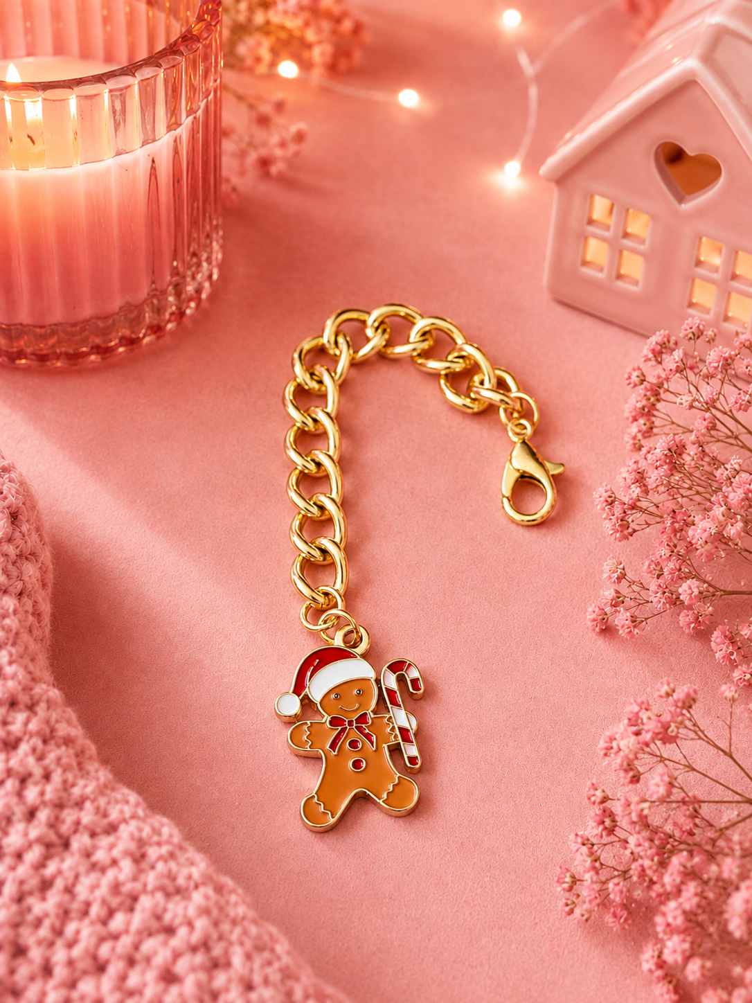 A gold-toned chain charm with a gingerbread man charm wearing a Santa hat and holding a candy cane, styled on a pink fabric surface with a candle, string lights, a small house ornament, and dried pink flowers.