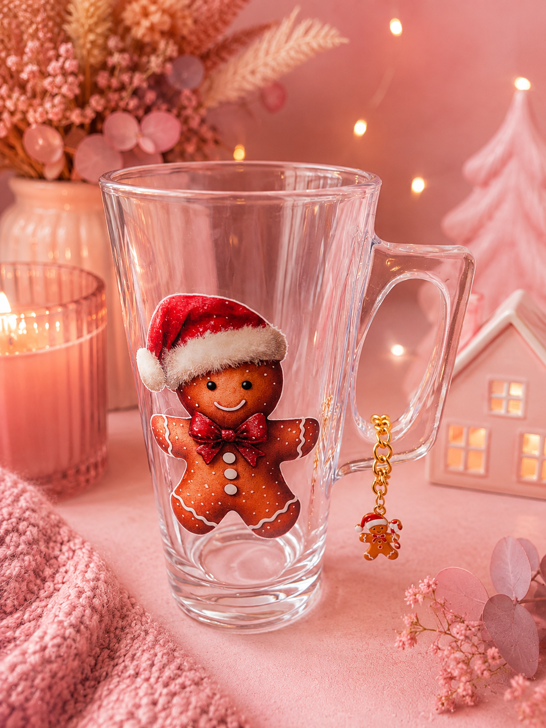 A clear glass mug with a handle, decorated with a gingerbread man wearing a red Santa hat and bow tie. The mug is styled on a pink surface with a pink knit blanket, a lit candle, dried flowers, and a small pink house in the background.