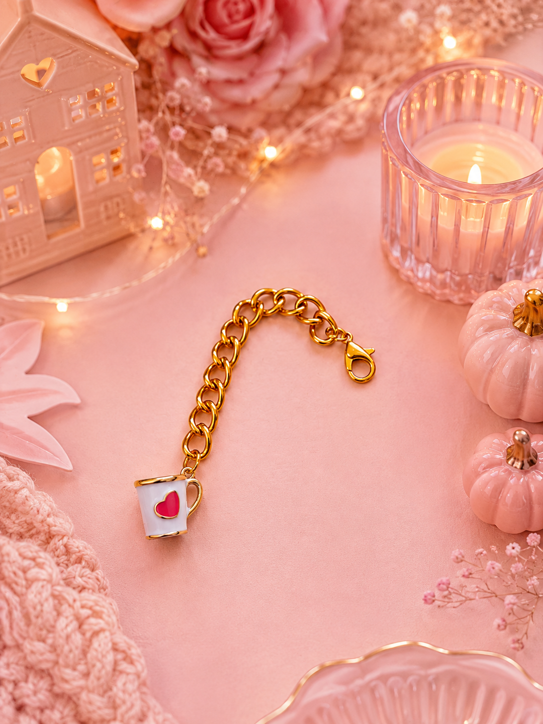 A gold chain charm with a white teacup charm featuring a pink heart, styled on a soft pink background with decorative props including a candle, small pumpkins, and a miniature house.