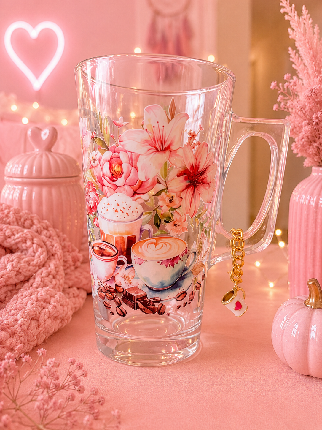 A clear glass mug with a floral and coffee-themed print, decorated with a gold heart charm, set against a soft pink background with decorative props.