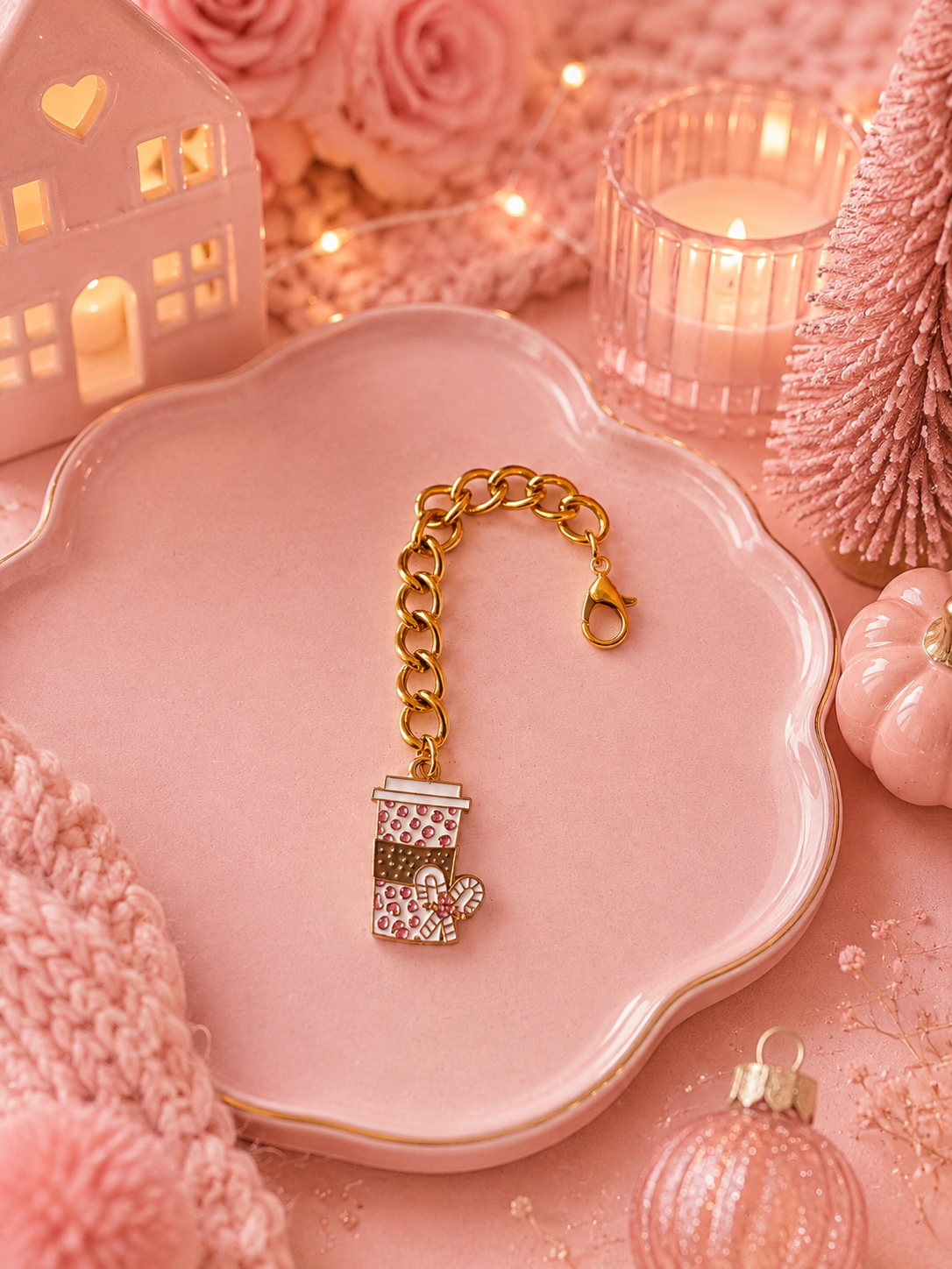 A gold-tone chain charm with a Christmas coffee cup charm covered in pink rhinestones, displayed on a scalloped pink tray surrounded by cozy holiday props including a candle, miniature house, and pink knit textures.