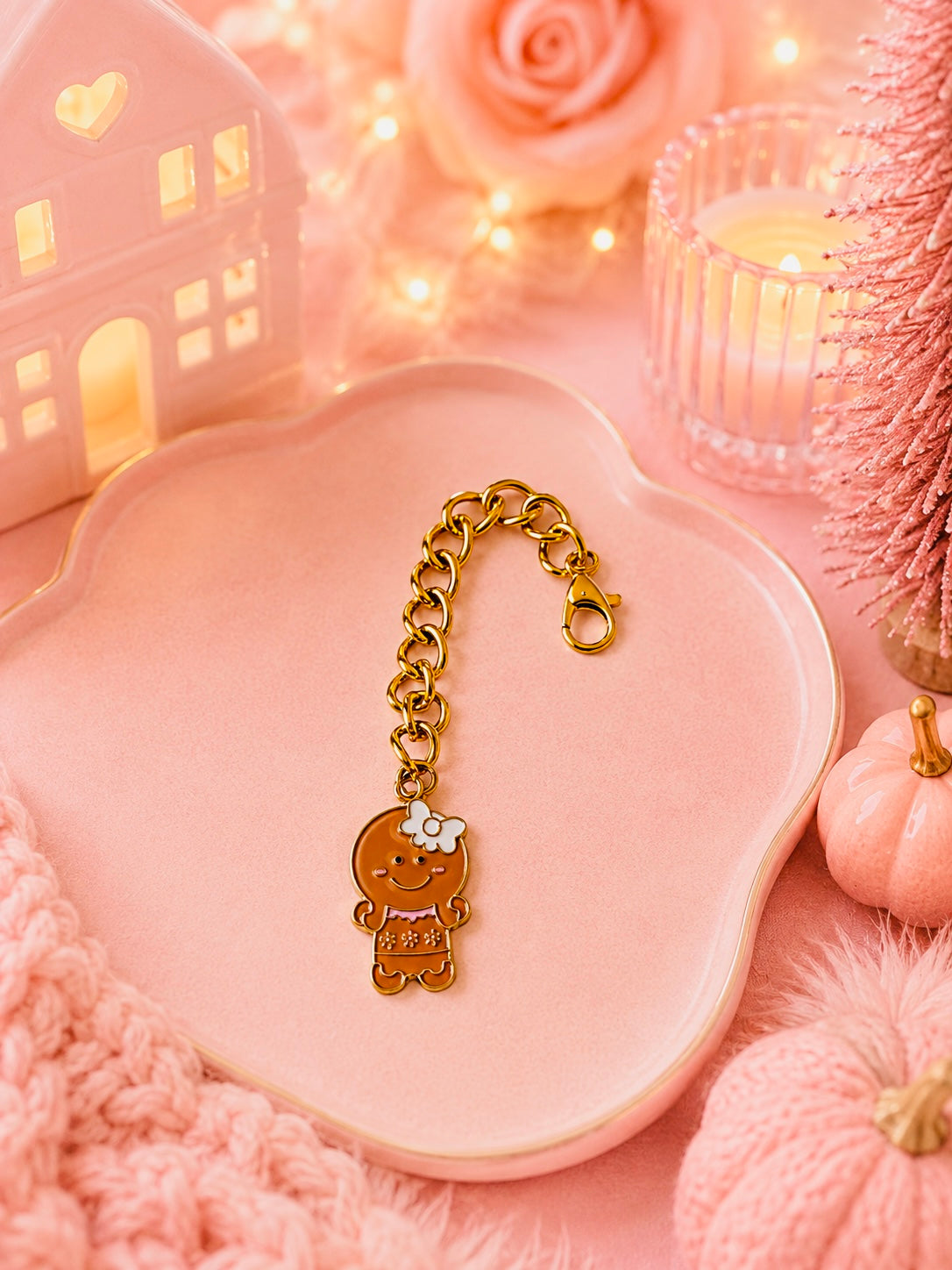A gold-tone charm with a gingerbread character pendant, displayed on a pink scalloped tray surrounded by cozy pink holiday decorations, including a candle, miniature tree, and warm lights.