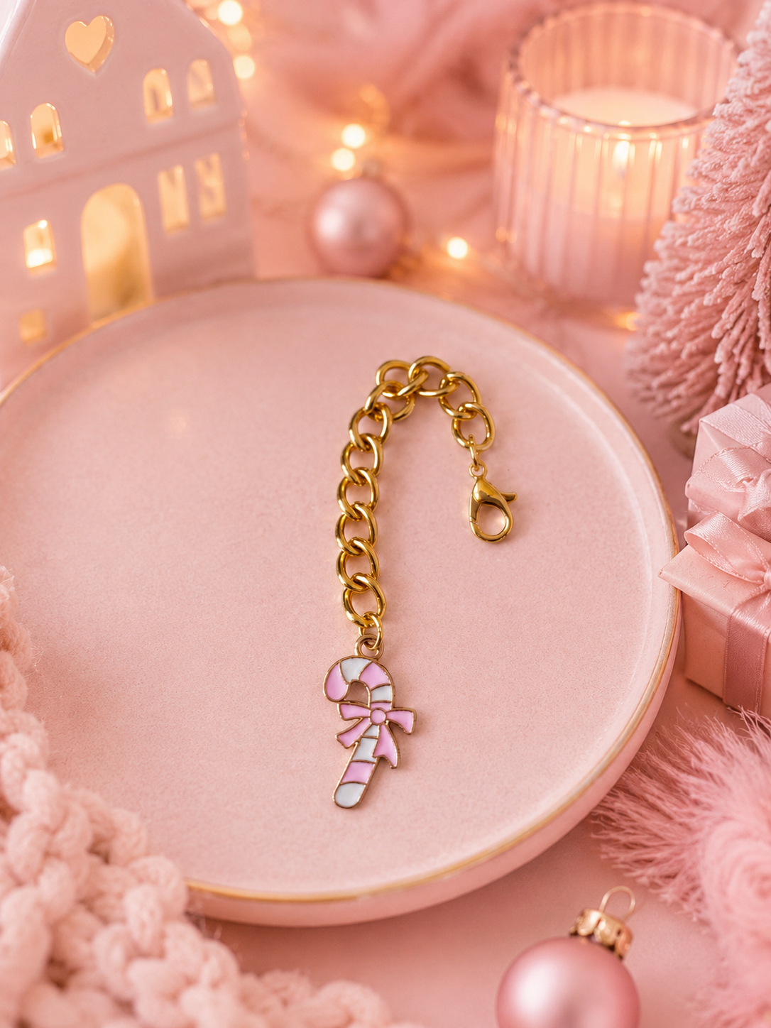 A gold-tone chain with a candy cane charm, styled on a pink ceramic dish with pink holiday decorations, a candle, and a gift box.