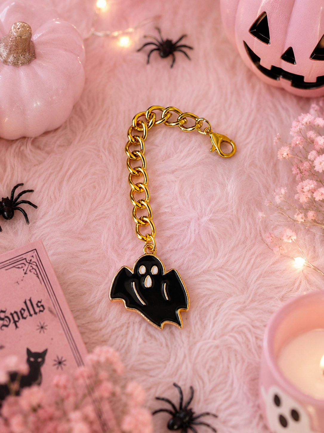 A gold chain with a black and white ghost charm, styled on a pink faux fur background with Halloween props including a jack-o'-lantern, small pumpkins, black spiders, fairy lights, and a pink book.
