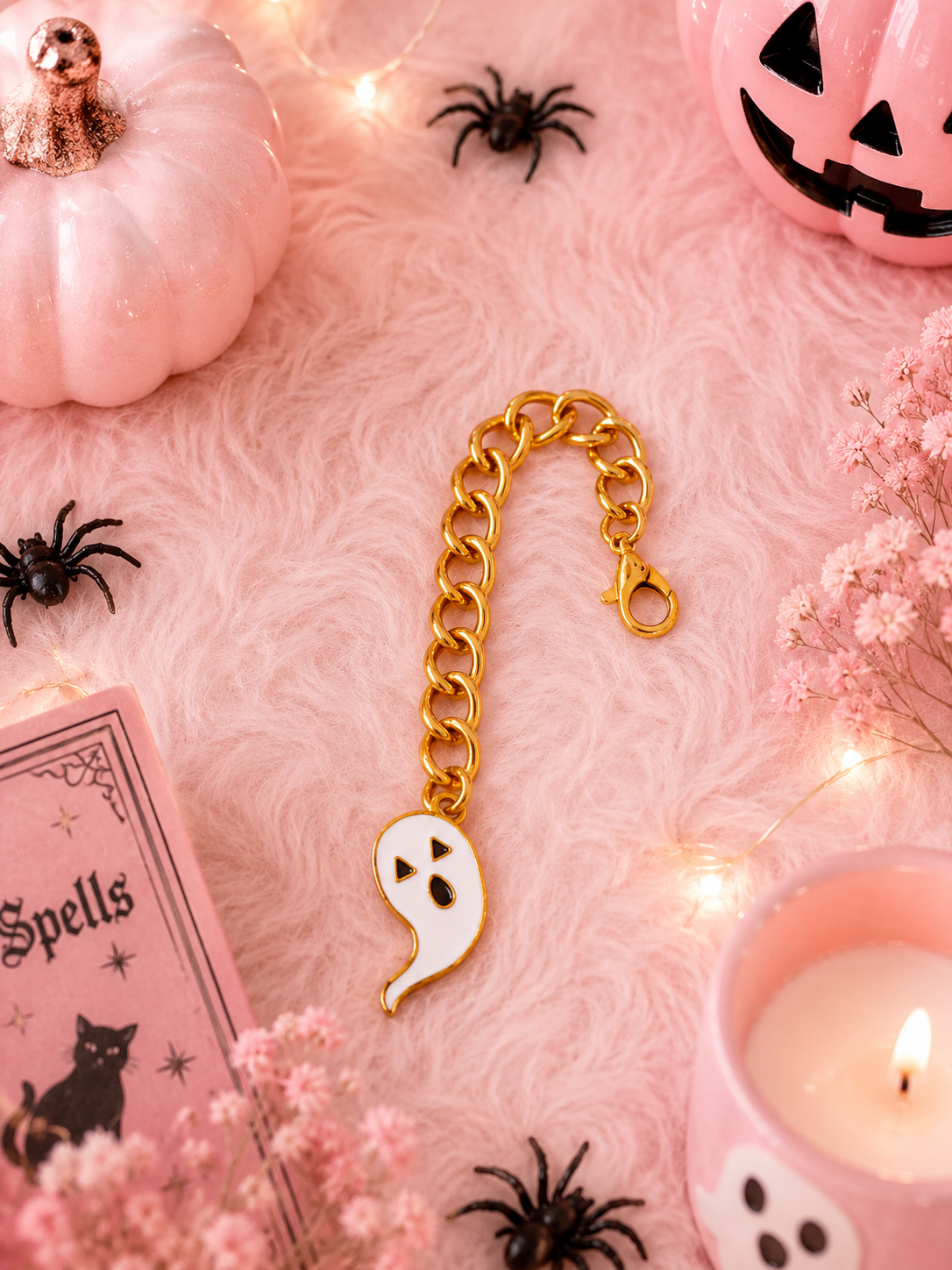 A gold chain with a white ghost charm, styled on a pink faux fur surface with Halloween props including a pink pumpkin, black spiders, a candle, and a book.