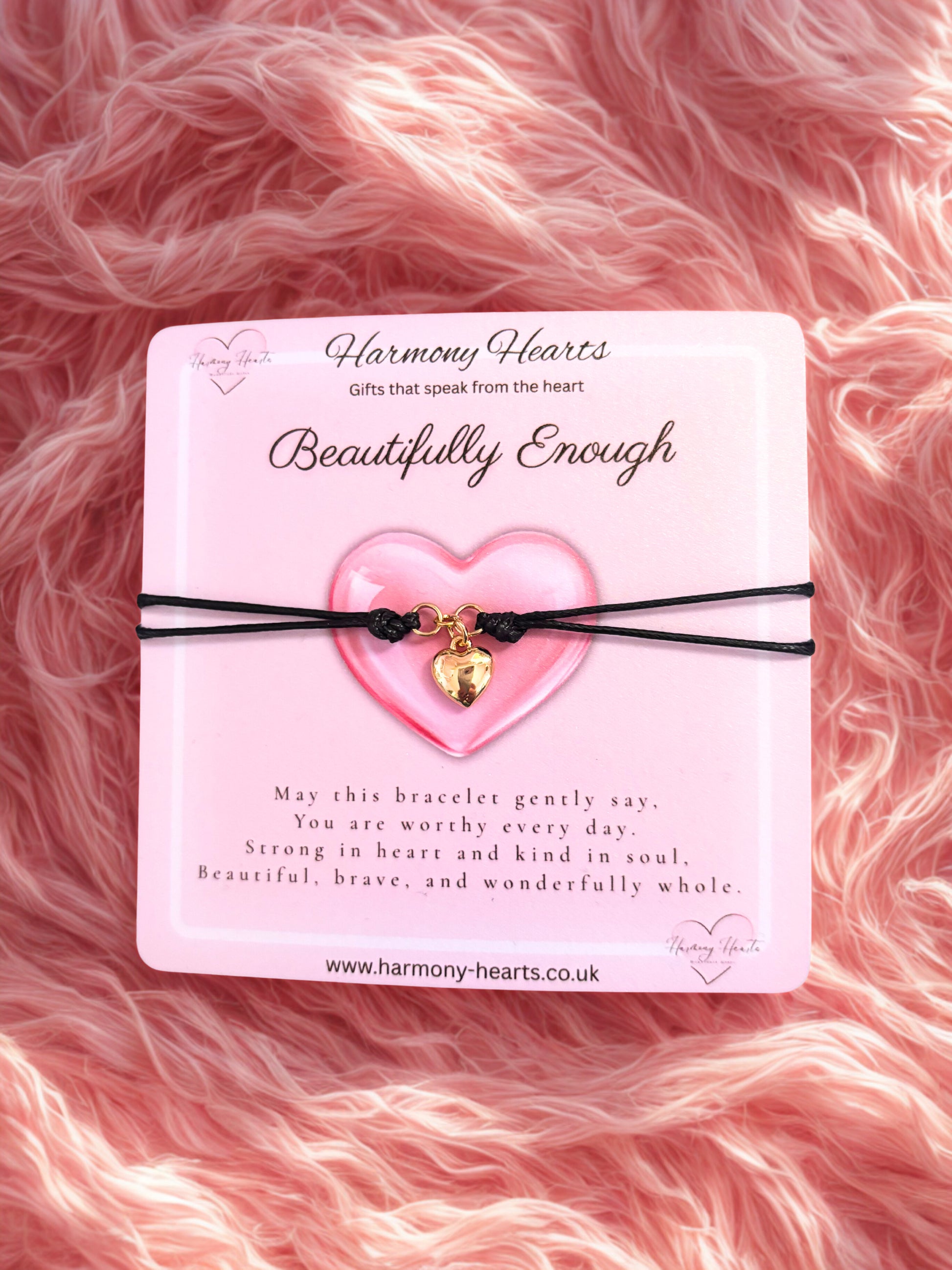 Heart-shaped bracelet with gold charm on a pink card with text, against a pink fluffy background