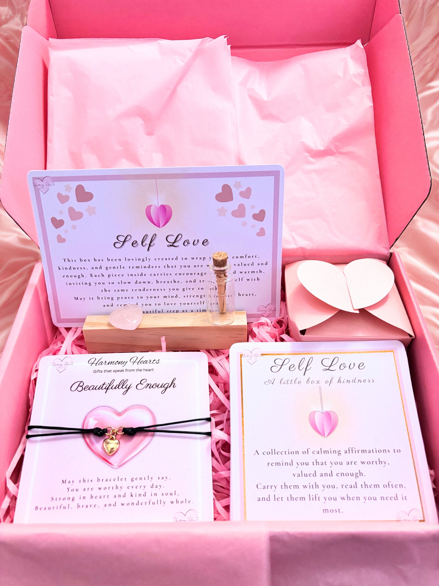Pink gift box with self-love cards, bracelet, and heart-shaped object on a pink background