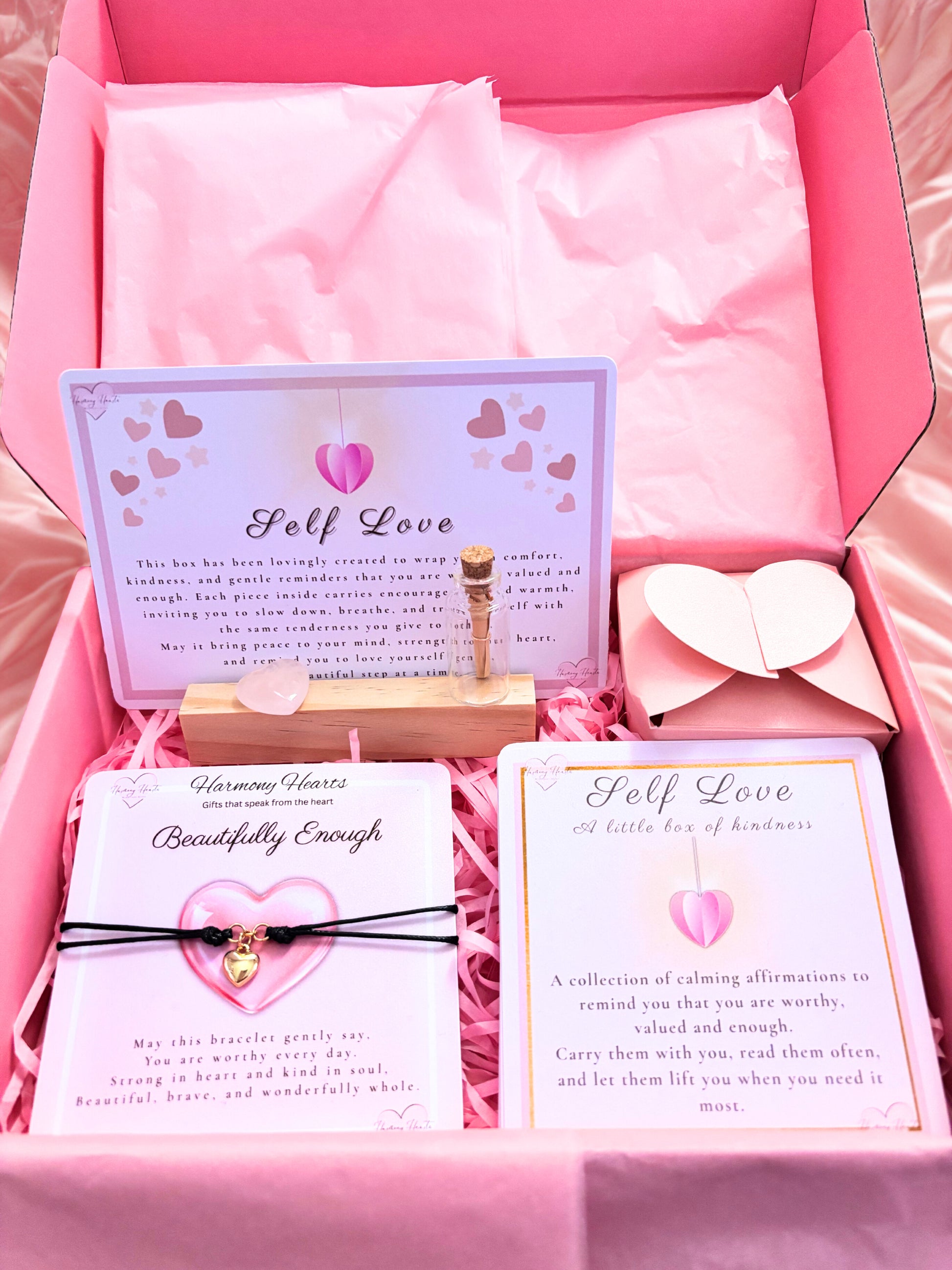 Pink gift box with self-love cards, bracelet, and heart-shaped object on a pink background