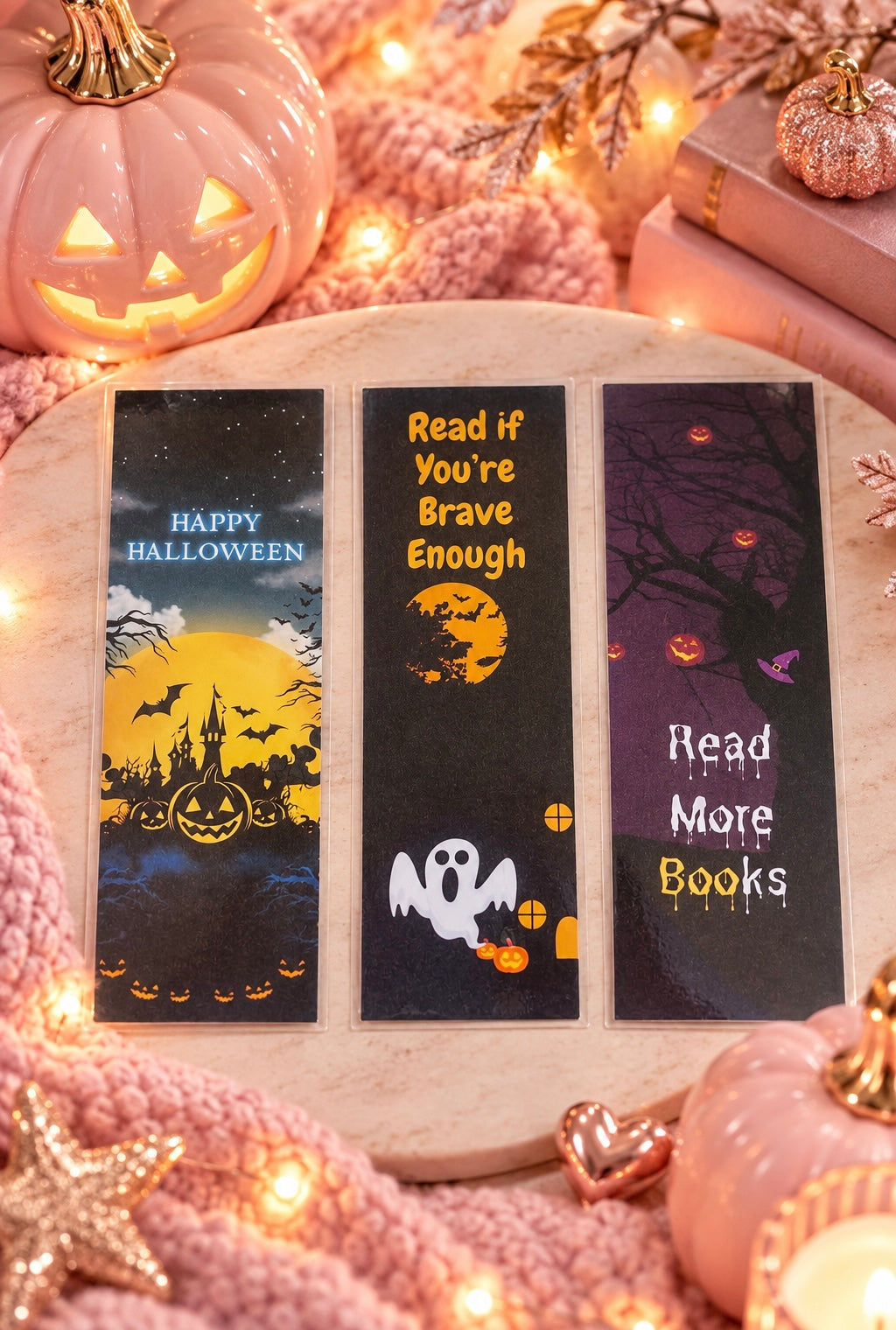 Three Halloween-themed book bookmarks displayed on a wooden tray, surrounded by cozy fall decor including a glowing jack-o'-lantern, pink knit fabric, candles, and stacked books.