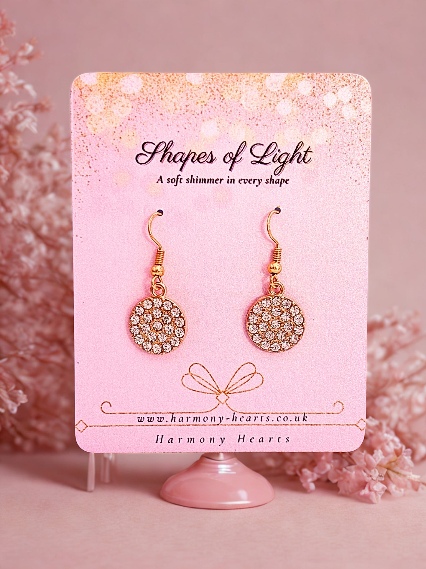 Pair of round earrings with gold and crystal details on a 'Shapes of Light' card with pink background.