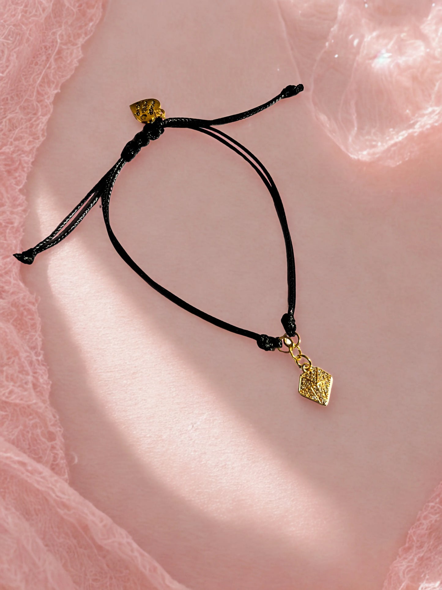 Black cord bracelet with gold diamond shaped charm on a pink textured background