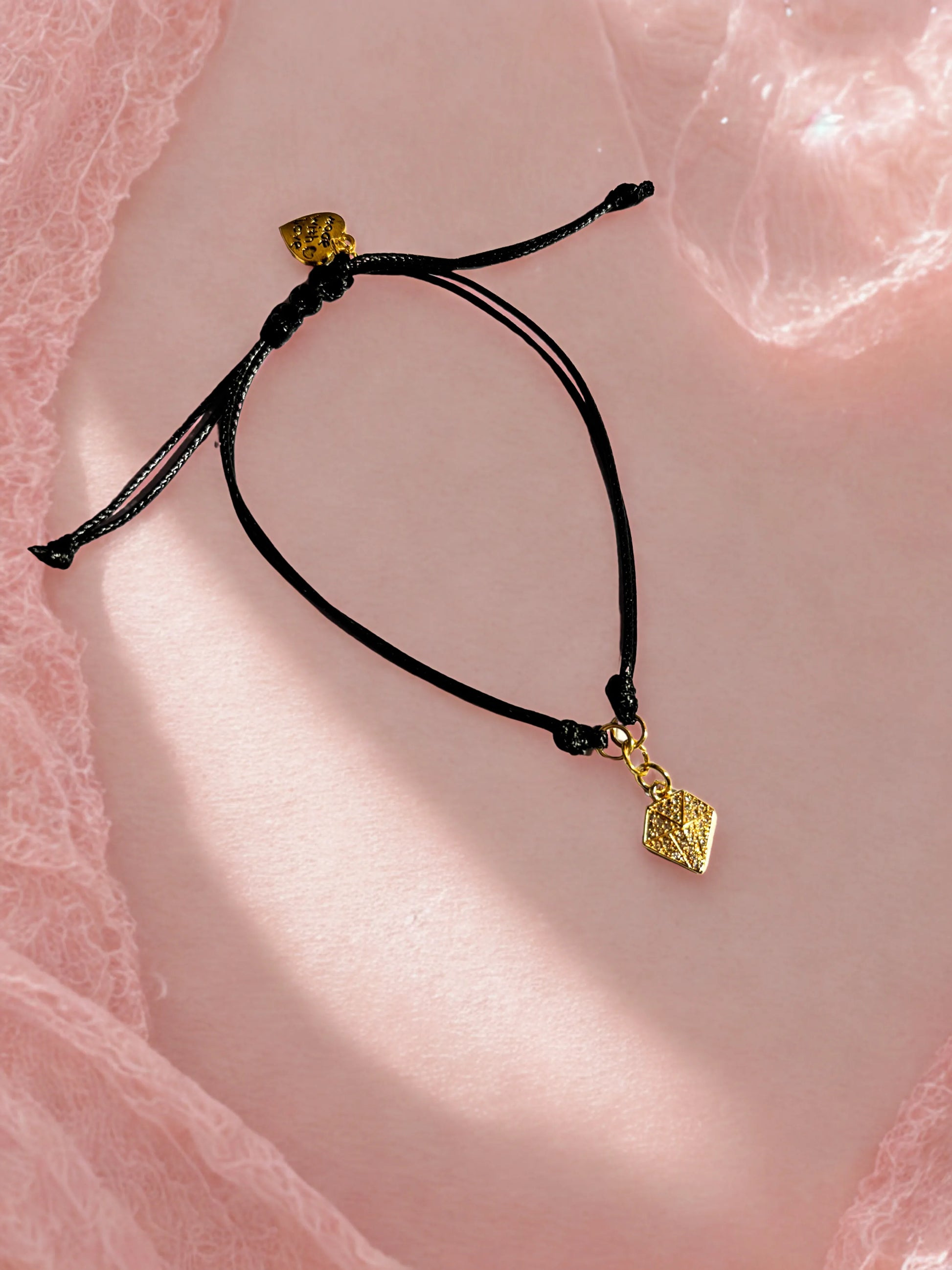 Black cord bracelet with gold diamond shaped charm on a pink textured background