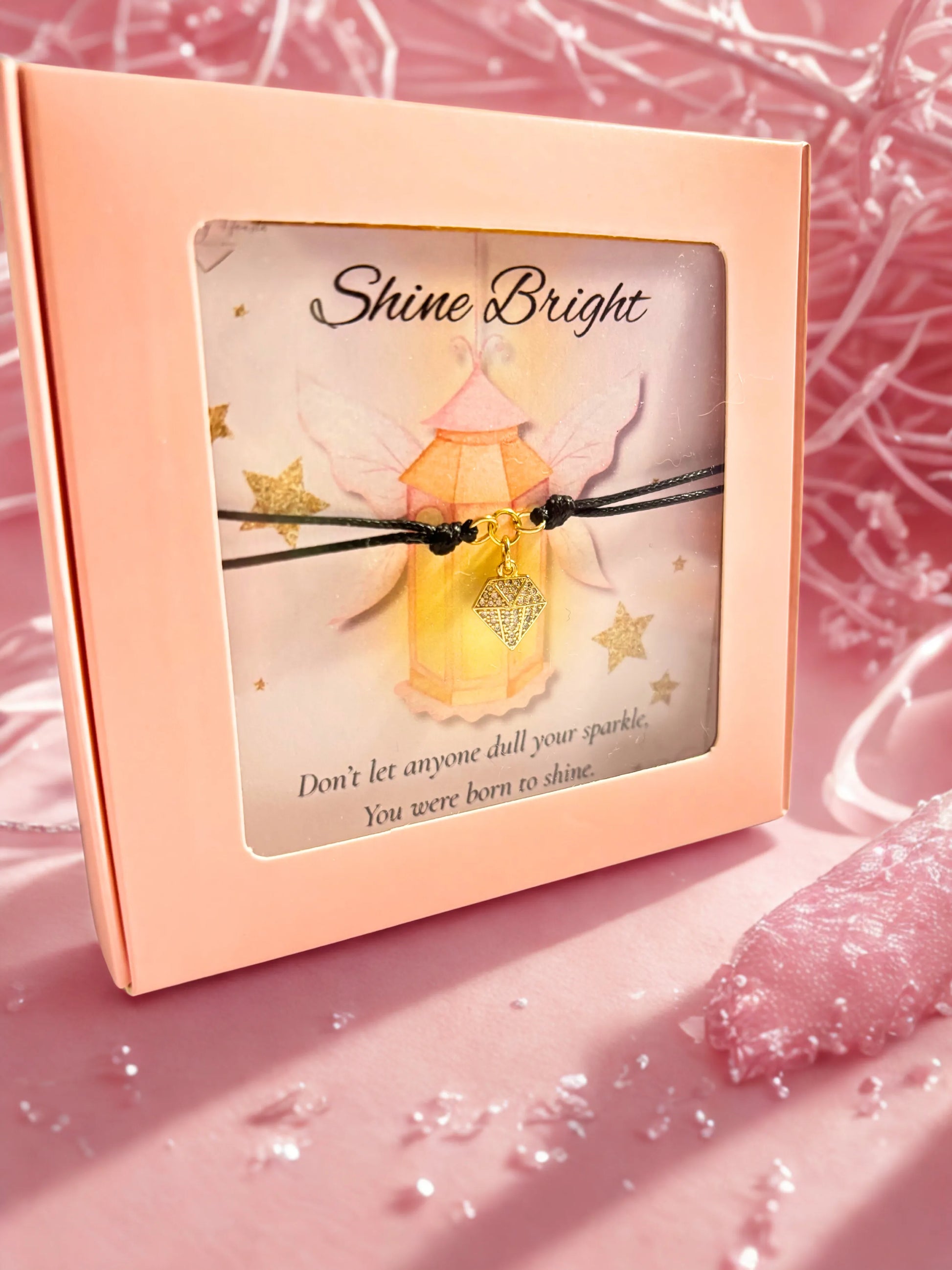 Bracelet with a gold diamond shaped charm in a pink box with 'Shine Bright' message on a pink background