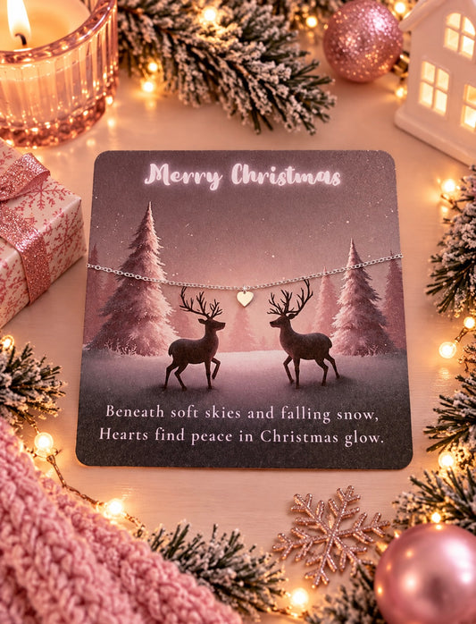 A Christmas-themed card featuring a silver heart bracelet with two deer silhouettes and winter trees, surrounded by holiday decorations including lights, pine branches, ornaments, and a candle.