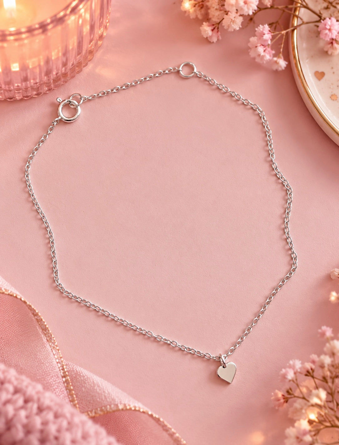 A delicate silver chain bracelet with a small heart-shaped pendant, displayed on a soft pink background with decorative pink flowers and ribbon.
