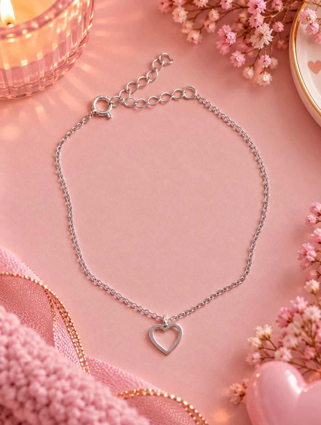 A silver chain bracelet with a small open heart pendant, displayed on a soft pink background with decorative pink flowers, a candle, and pink ribbon.
