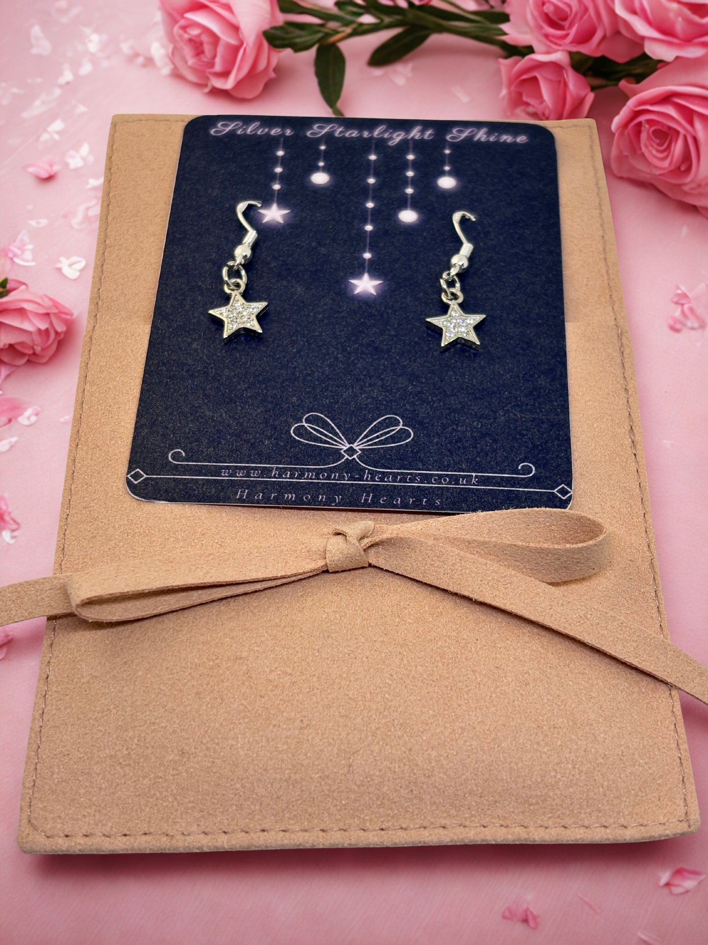 Star-shaped earrings on a card with a bow, surrounded by pink roses on a pink background