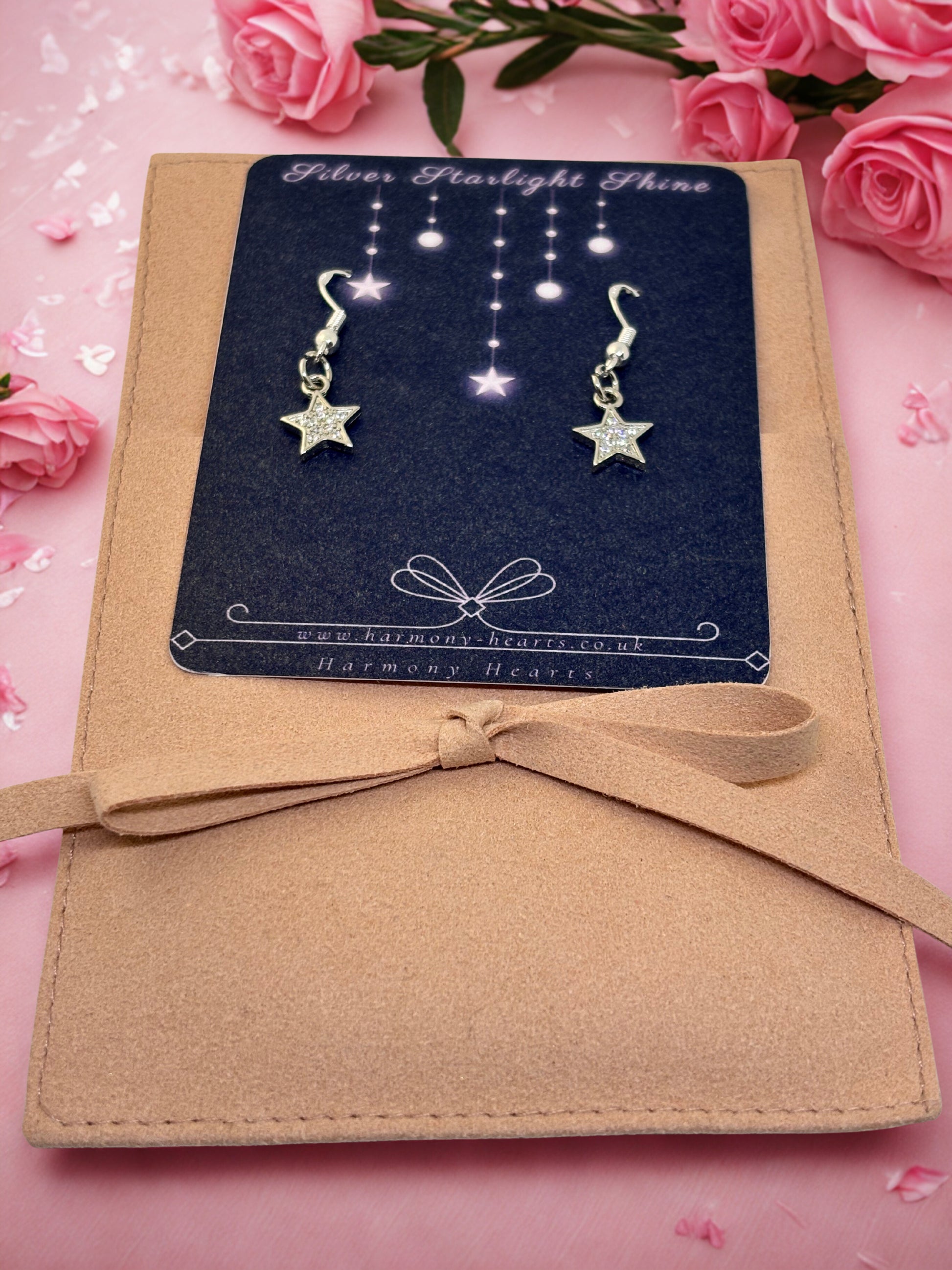 Star-shaped earrings on a card with a bow, surrounded by pink roses on a pink background