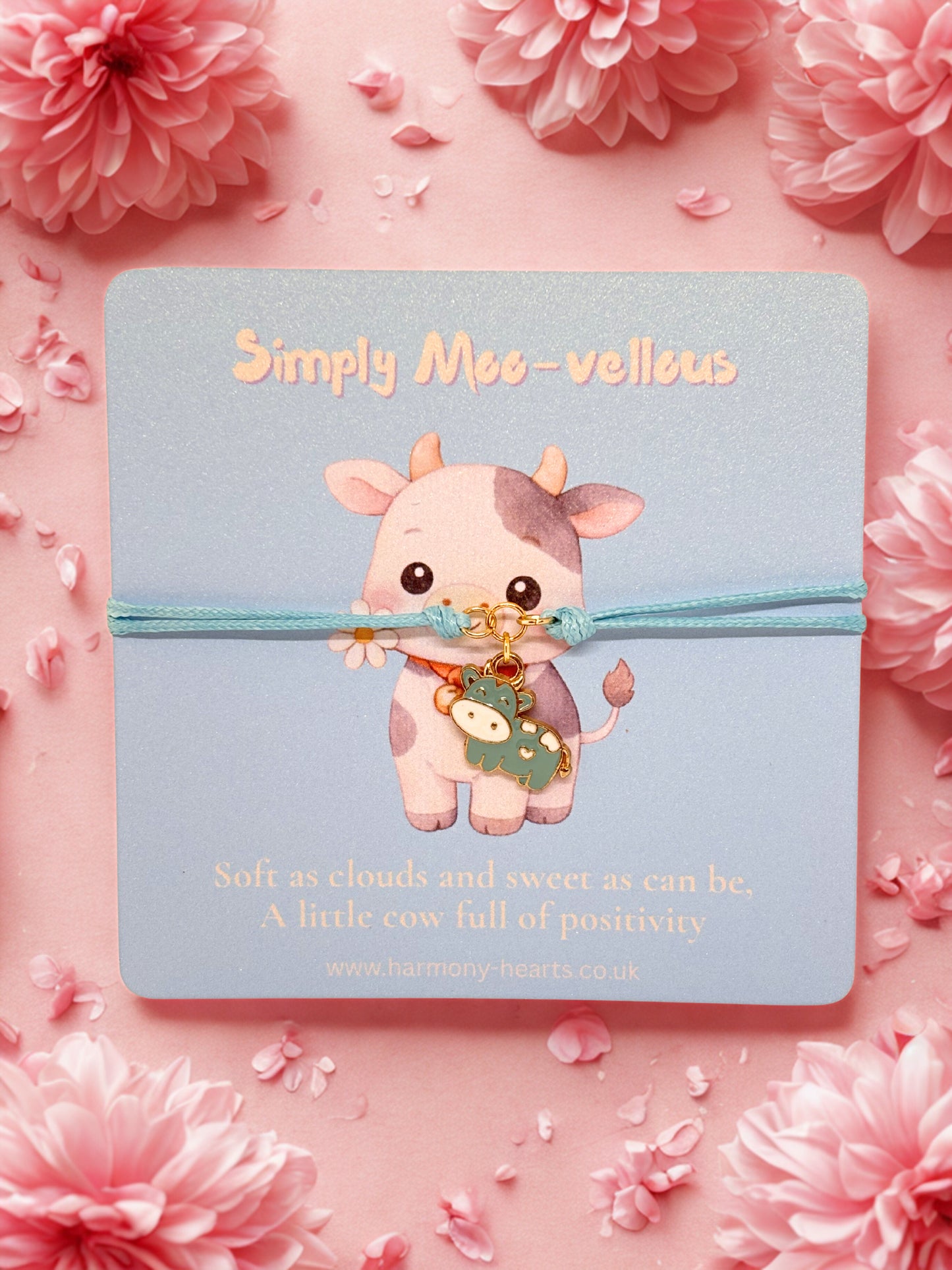 Blue cord bracelet with cow charm on a card labeled 'Simply Moo-vellous' surrounded by pink flowers.