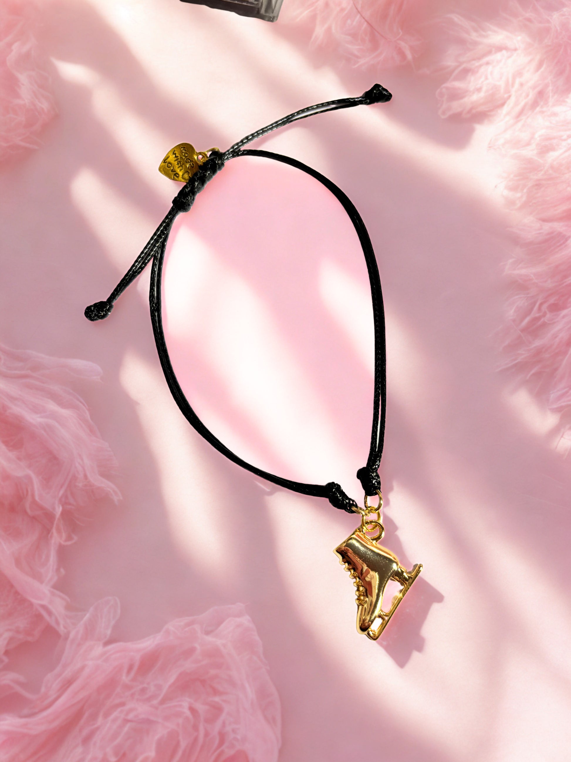 Black cord bracelet with a gold ice skate charm on a pink textured background