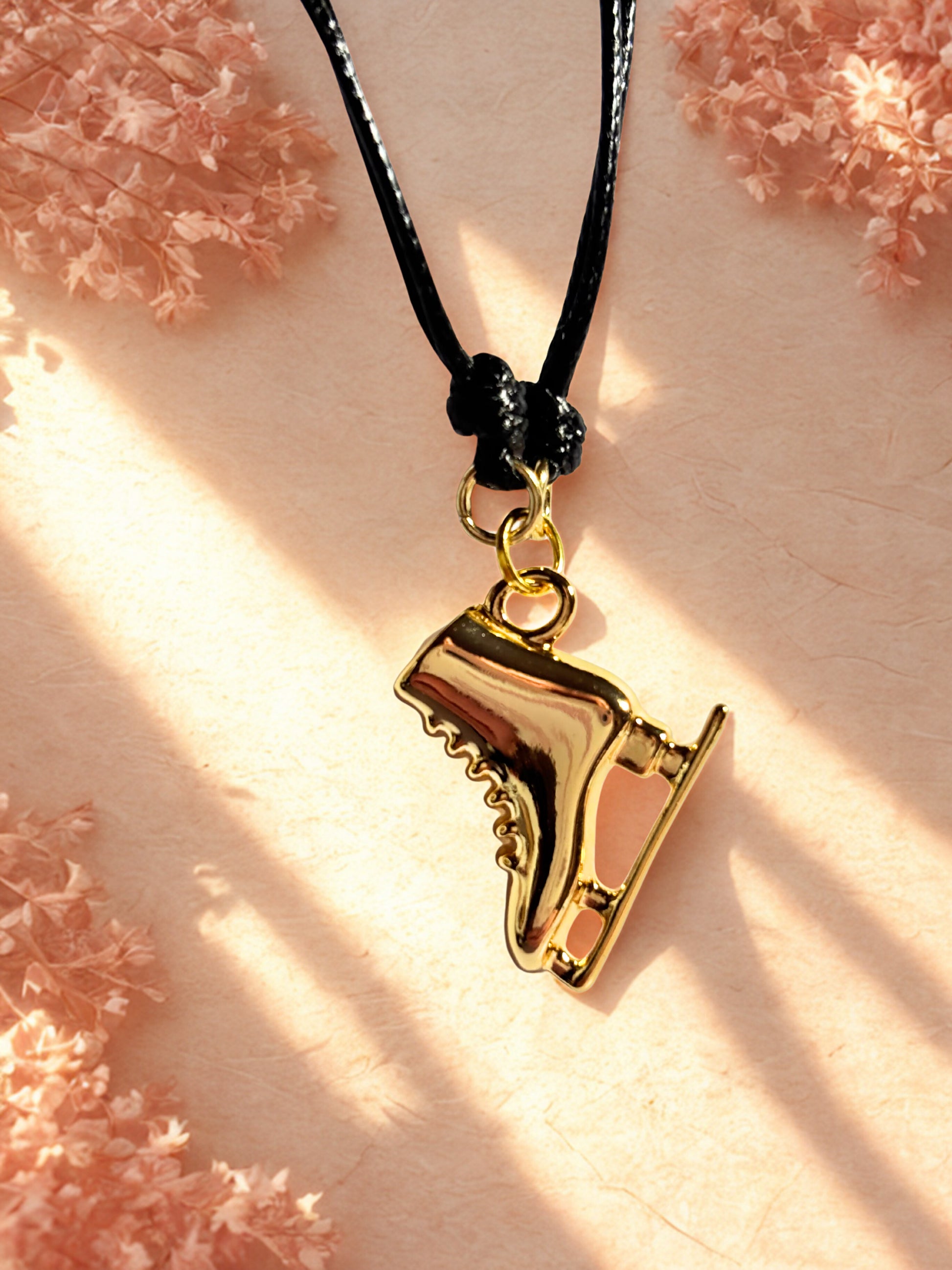 Gold ice skate pendant on a black cord against a soft pink floral background