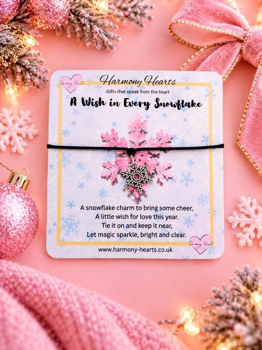 A pink snowflake charm wrapped in a black string and presented on a branded gift card from Harmony Hearts, styled on a pink background with holiday decorations including ornaments, lights, and a ribbon.