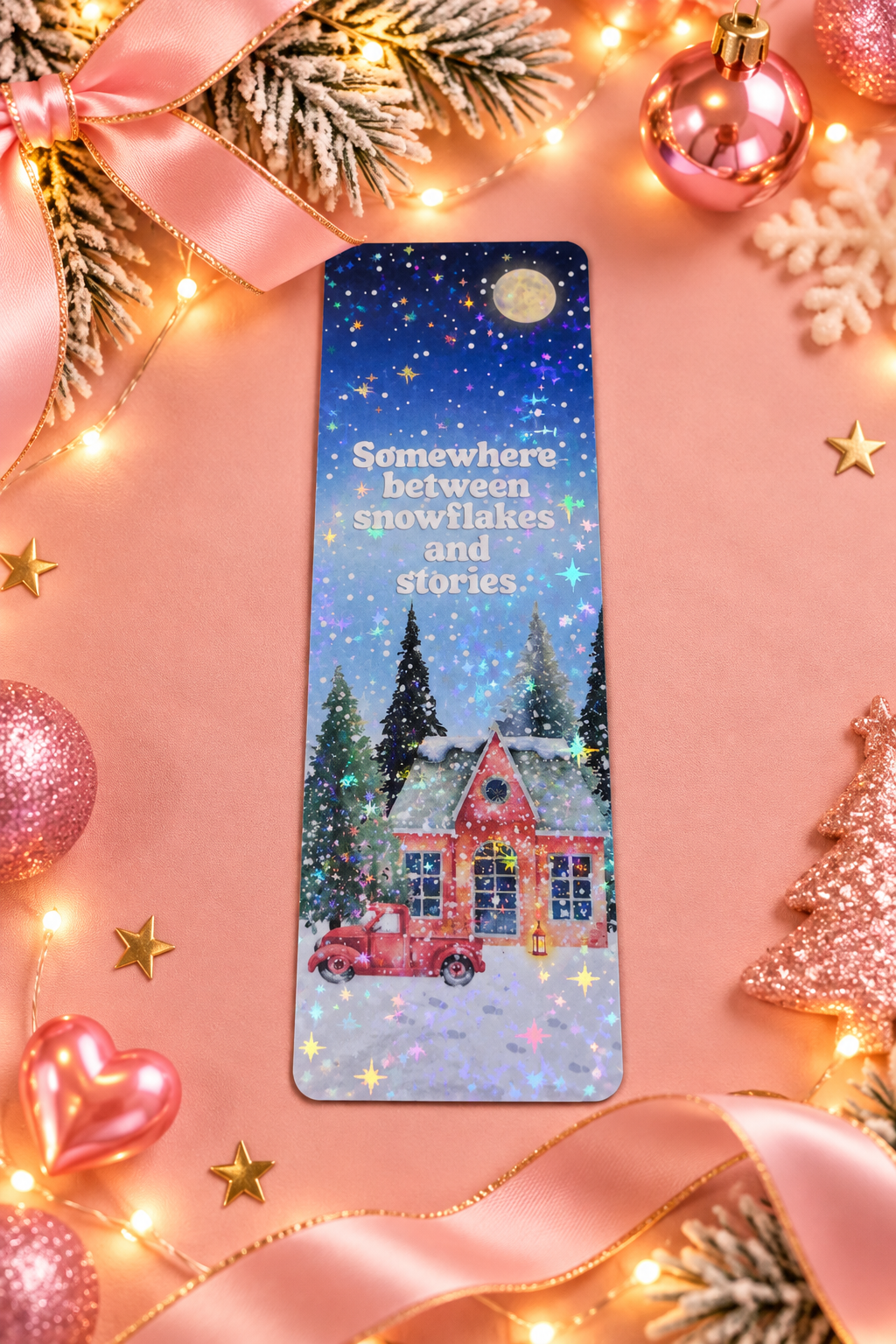 A rectangular printed bookmark with a glittery winter village scene and the phrase 'Somewhere between snowflakes and stories.' is centered on a pink holiday-themed background with ribbon, ornaments, lights, and stars.