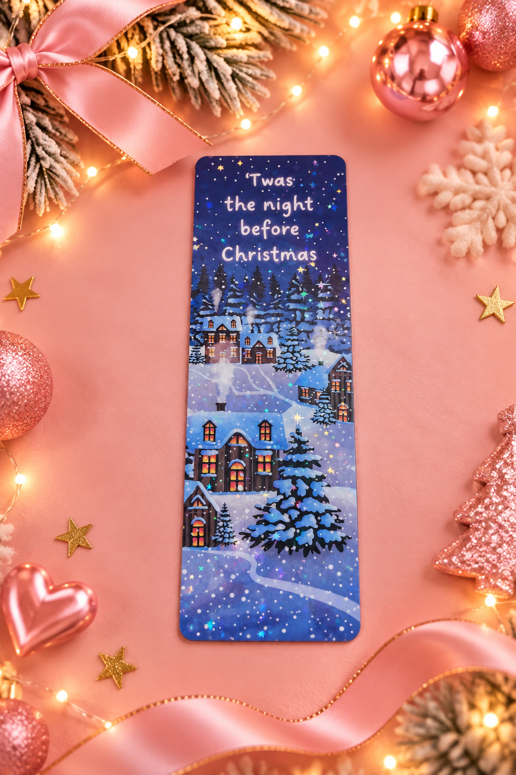 A rectangular Christmas-themed bookmark with the text 'Twas the night before Christmas' and an illustrated snowy village scene, placed on a pink background with festive decorations including lights, ornaments, ribbon, and snowflake accents.