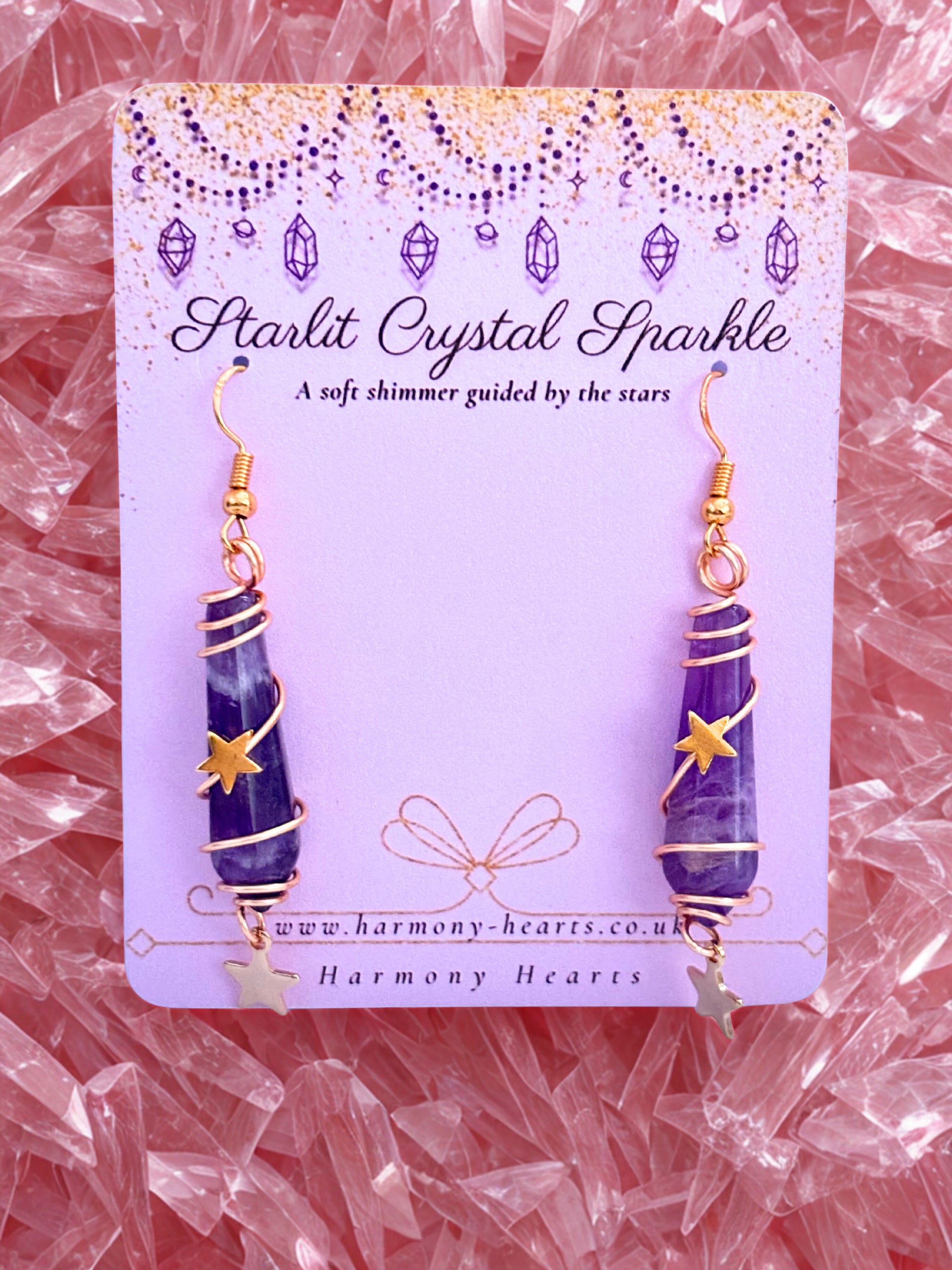 Purple amethyst crystal earrings on a 'Starlit Crystal Sparkle' card with gold accents, placed on a pink crystal background.