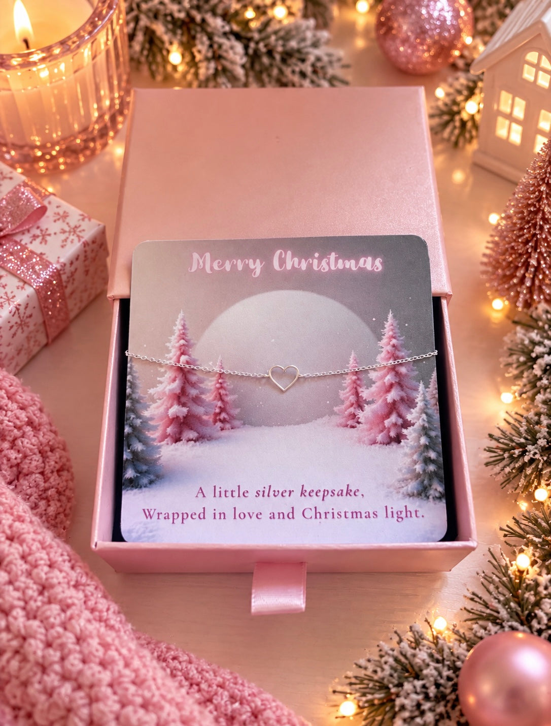 A silver keepsake bracelet with a heart-shaped link is displayed on a printed gift card inside a pink box. The card reads 'Merry Christmas' and 'A little silver keepsake.