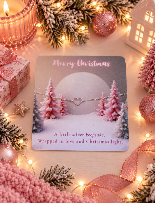 A delicate silver heart bracelet displayed on a printed Christmas-themed gift card, surrounded by festive holiday decorations including pink ornaments, pine branches, warm string lights, and wrapped gift boxes.