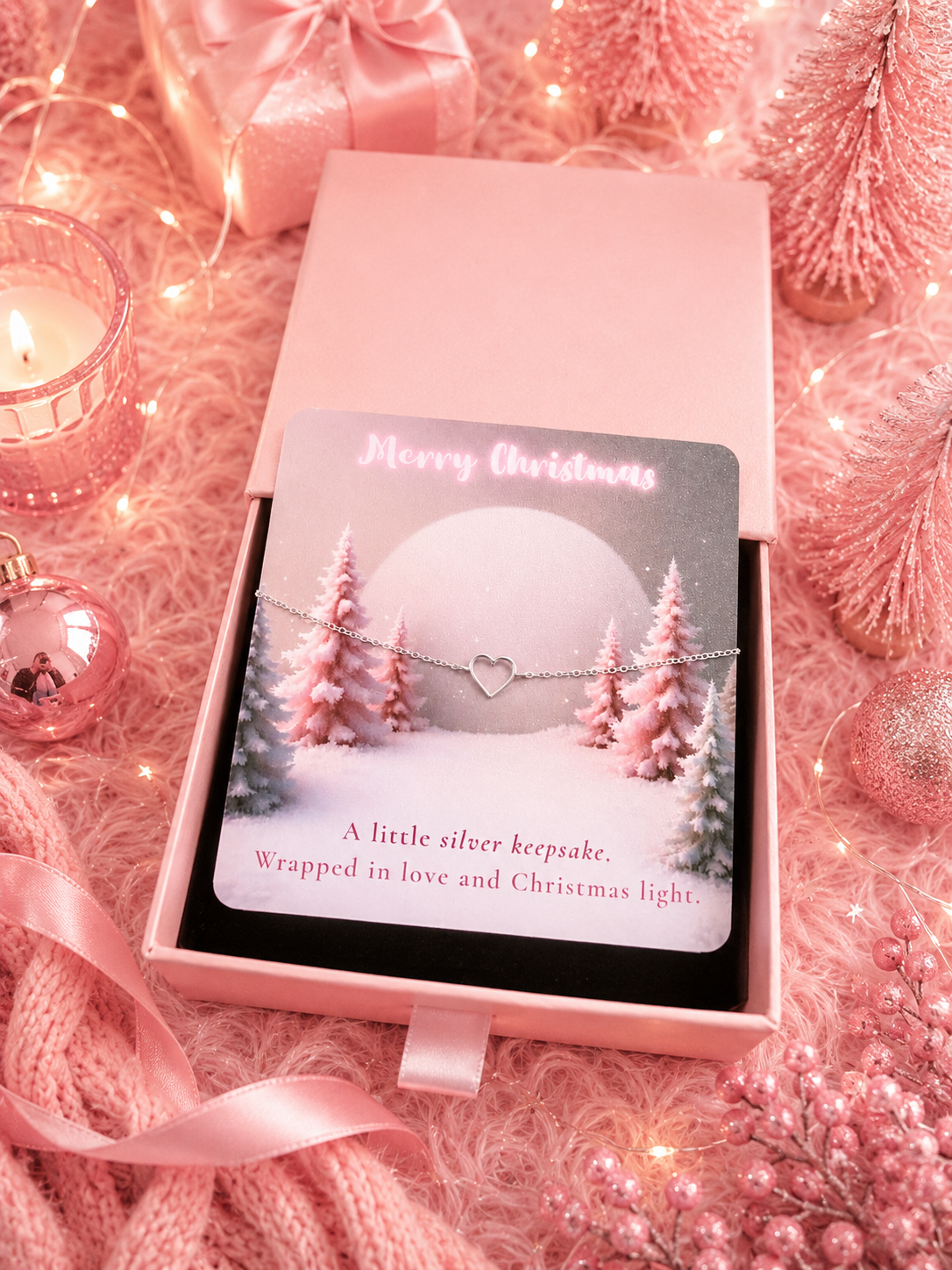 A silver keepsake bracelet with a heart charm is wrapped in a pink gift box with a Christmas-themed card. The box is surrounded by festive pink decorations, including a candle, ornaments, and fairy lights.