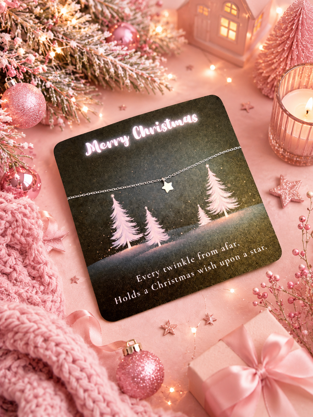 A silver bracelet with a star charm on a display card, styled in a cozy holiday flat-lay with pink ornaments, a knitted scarf, a candle, and a gift box.
