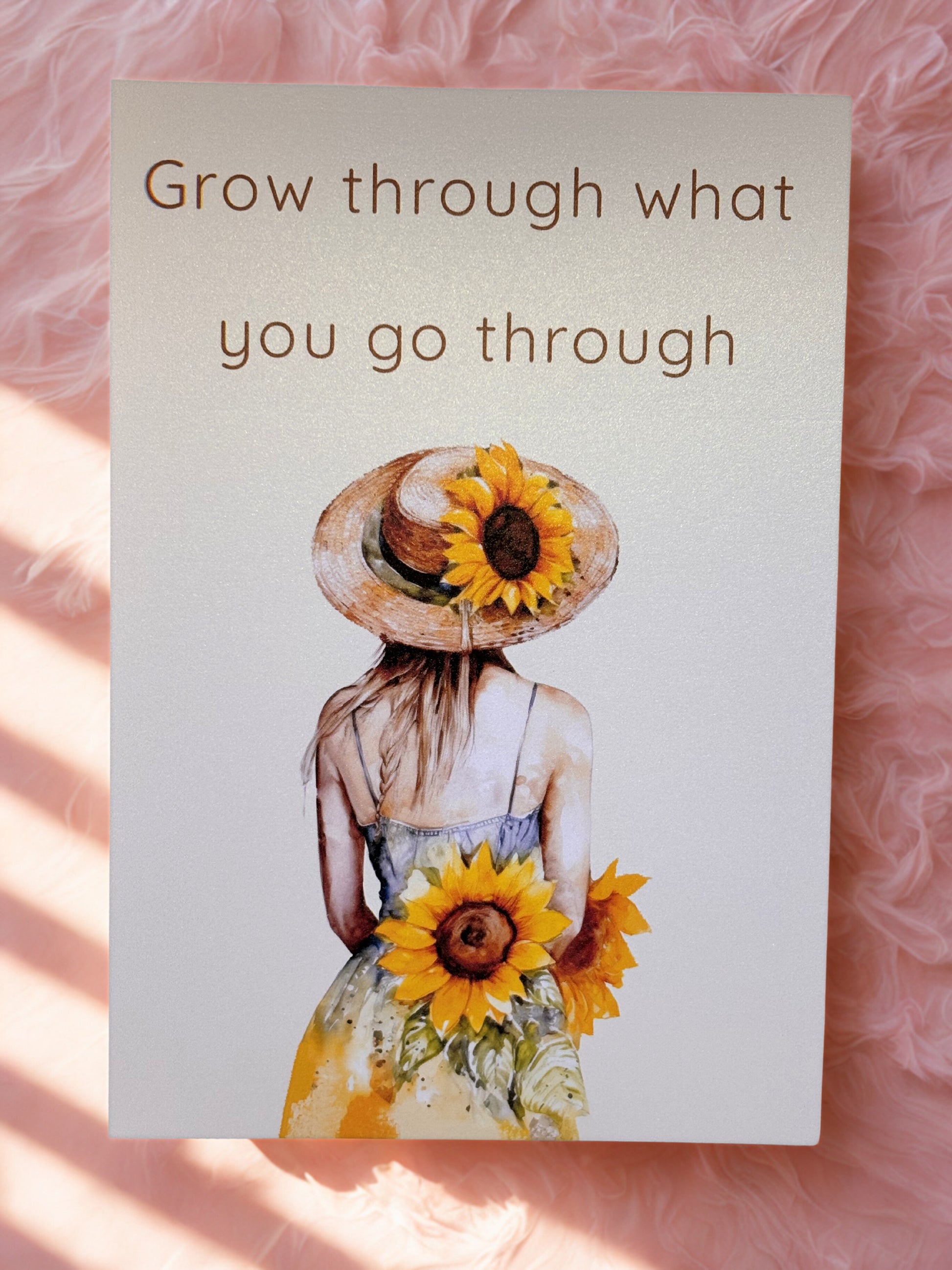 Motivational cardwith a woman wearing a sunflower hat and holding sunflowers, text 'Grow through what you go through' on a pink textured background.