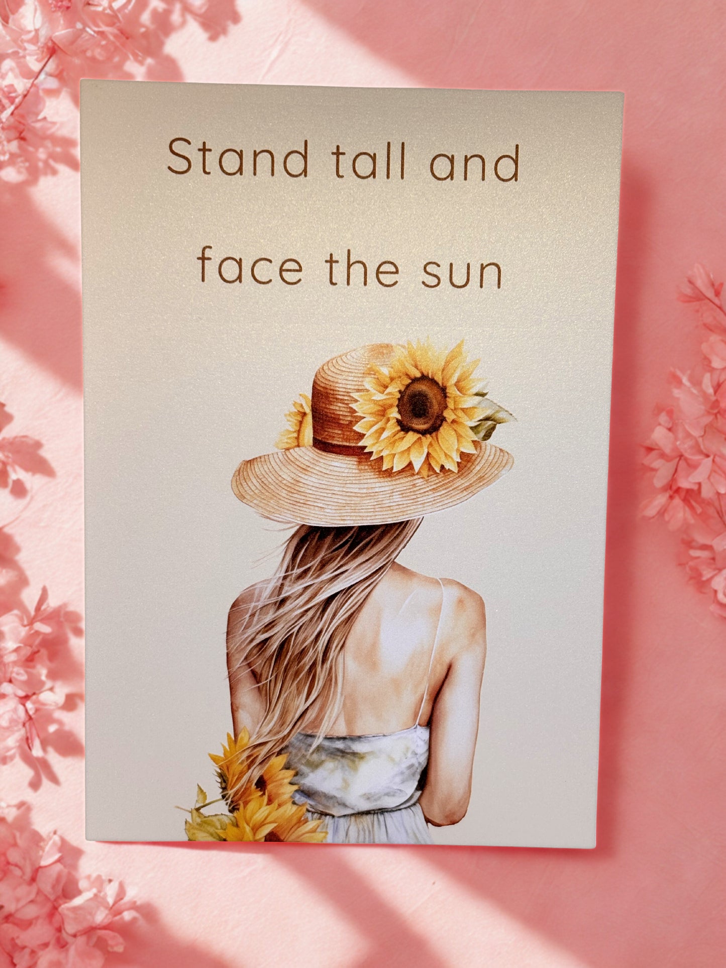 card with a woman wearing a sun hat with a sunflower, text 'Stand tall and face the sun', on a pink floral background.