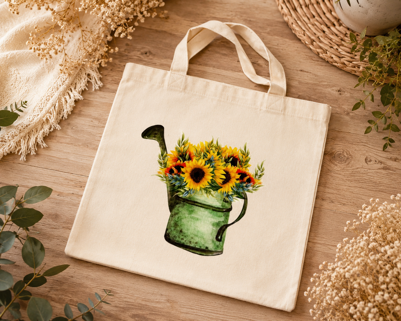 A cream canvas tote bag with a watercolor-style print of a green watering can holding bright yellow sunflowers, displayed on a wooden surface with dried flowers and greenery.