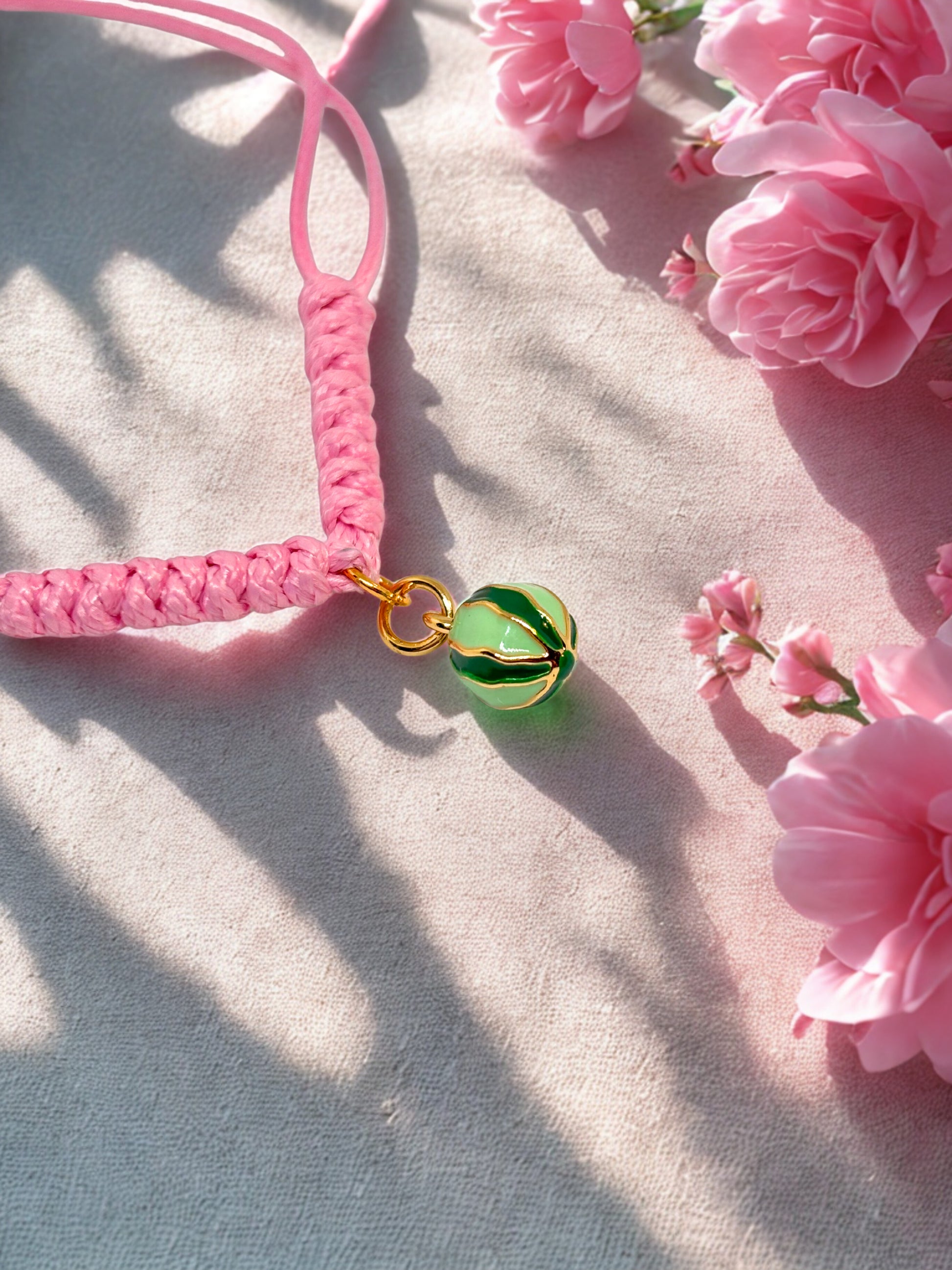 Pink braided bracelet with a green and gold watermelon charm on a light surface with pink flowers.