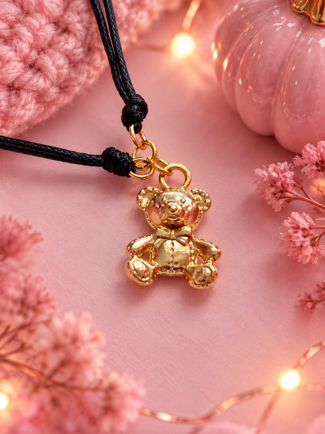 A gold teddy bear charm on a black cord bracelet, styled on a pink background with decorative pink flowers, a pink crocheted item, and warm fairy lights.
