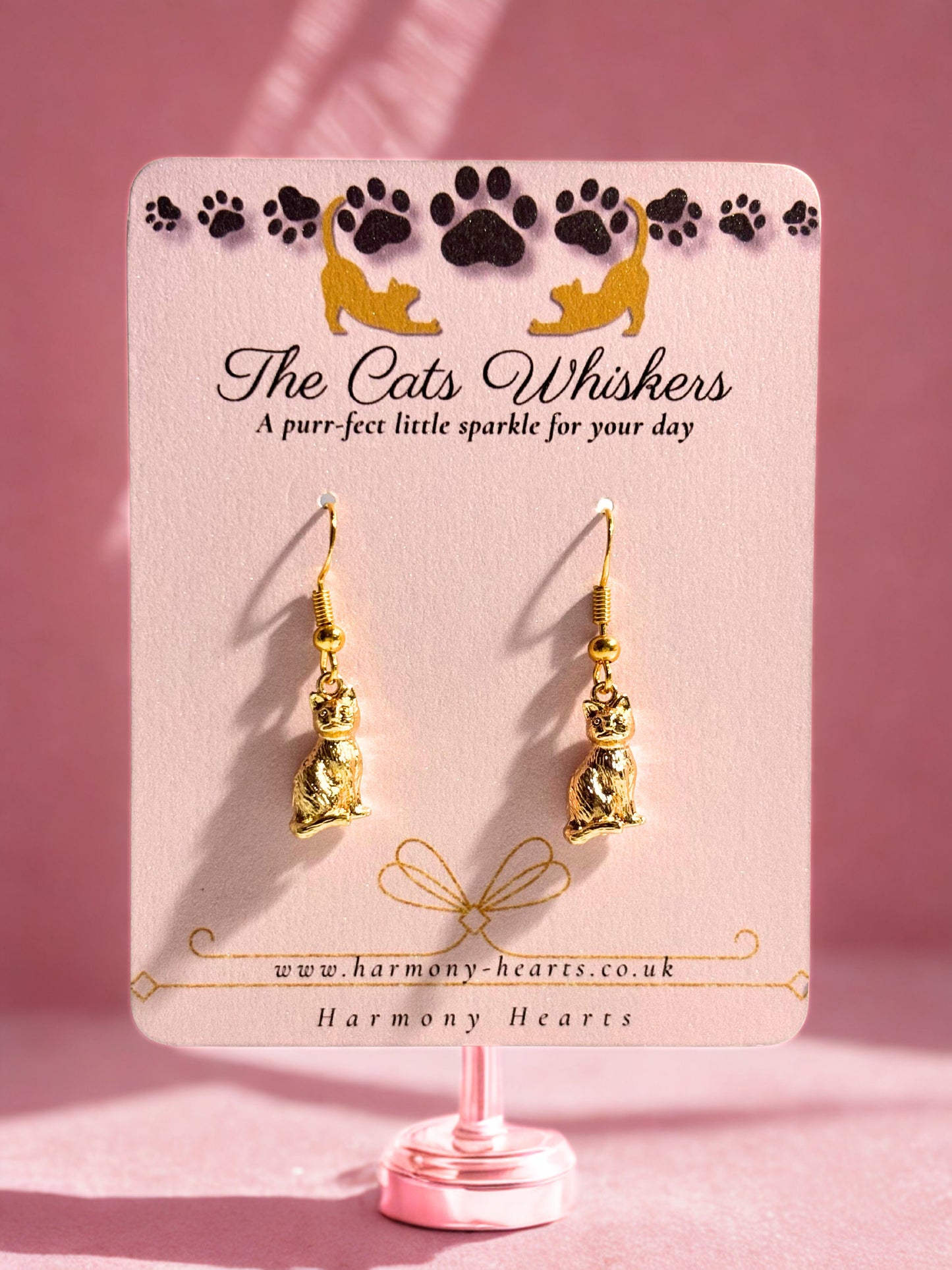 Gold cat-shaped earrings on a 'The Cats Whiskers' branded card with a pink background.