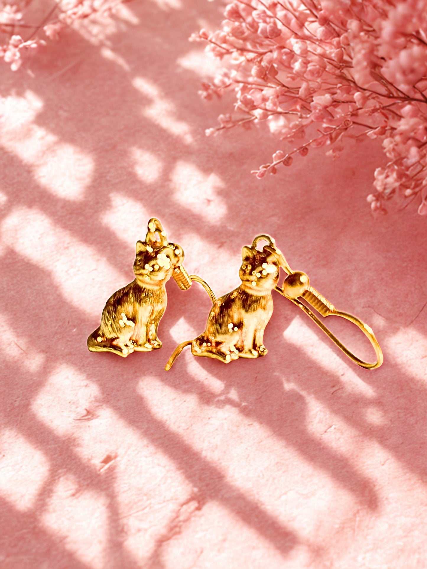 Gold cat-shaped earrings on a pink background with floral elements