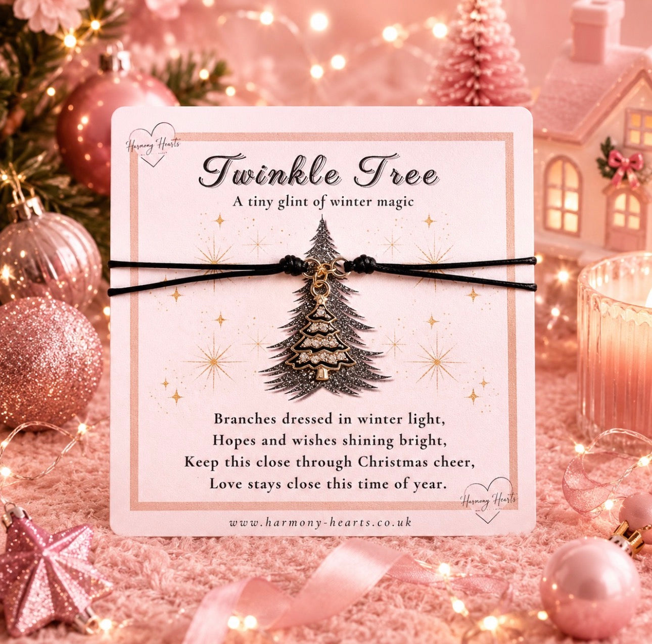 A square card reading 'Twinkle Tree' with a black cord bracelet featuring a small metallic Christmas tree charm, styled in a warm pink holiday setting with ornaments, lights, and a candle.
