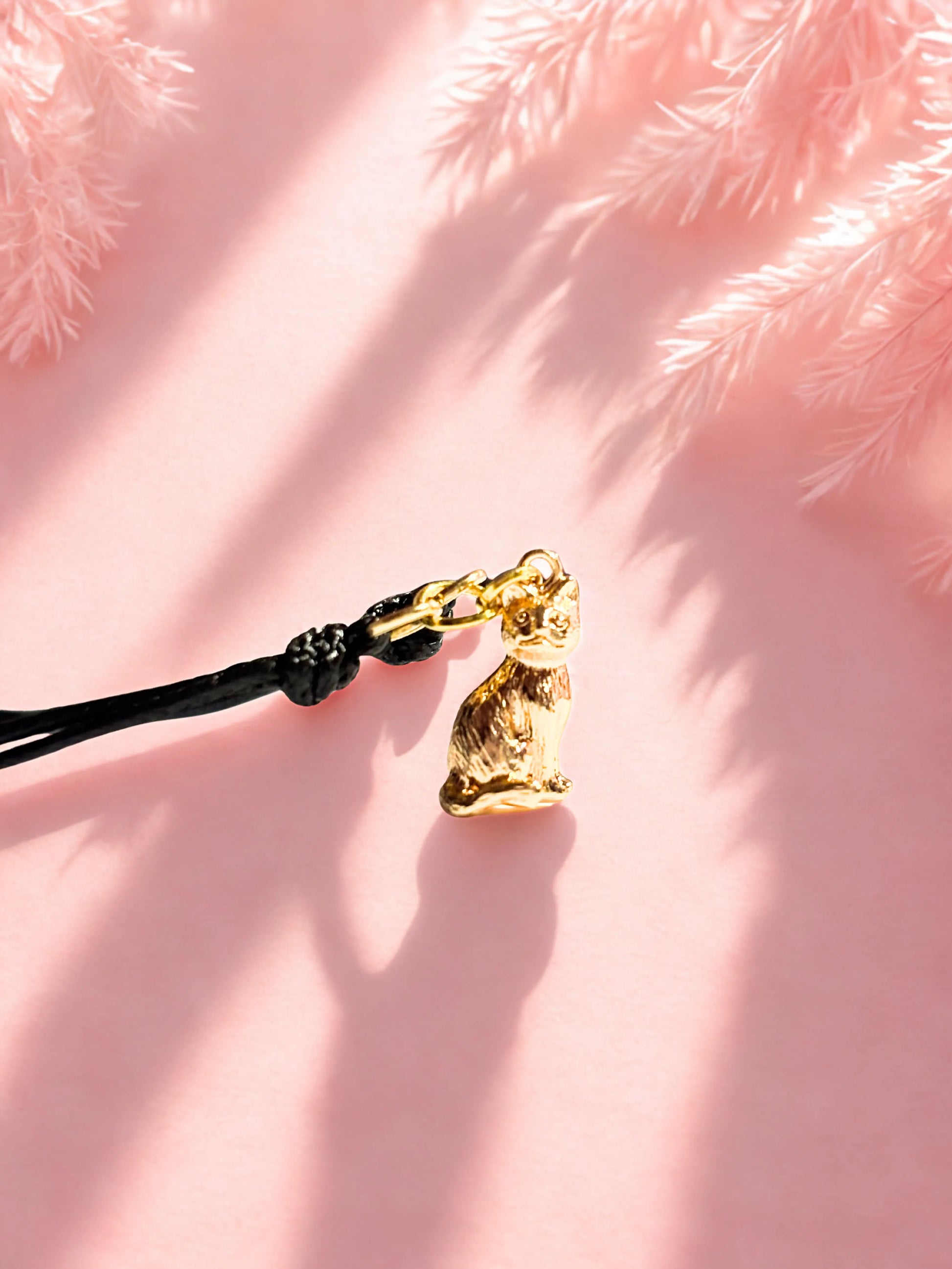 Gold cat charm on a black cord bracelet against a pink background