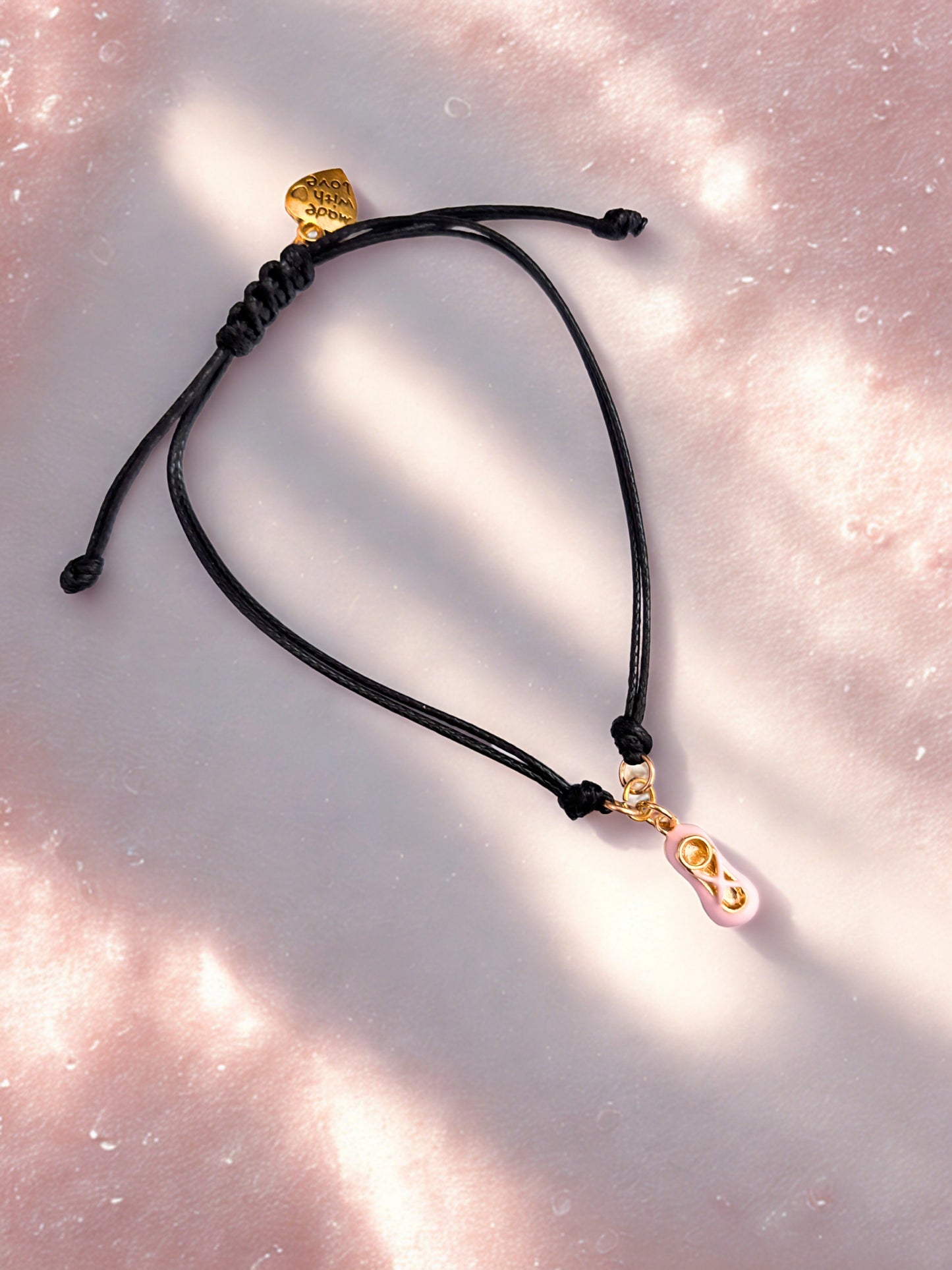 Black cord bracelet with gold ballet shoe charm on a pink textured background